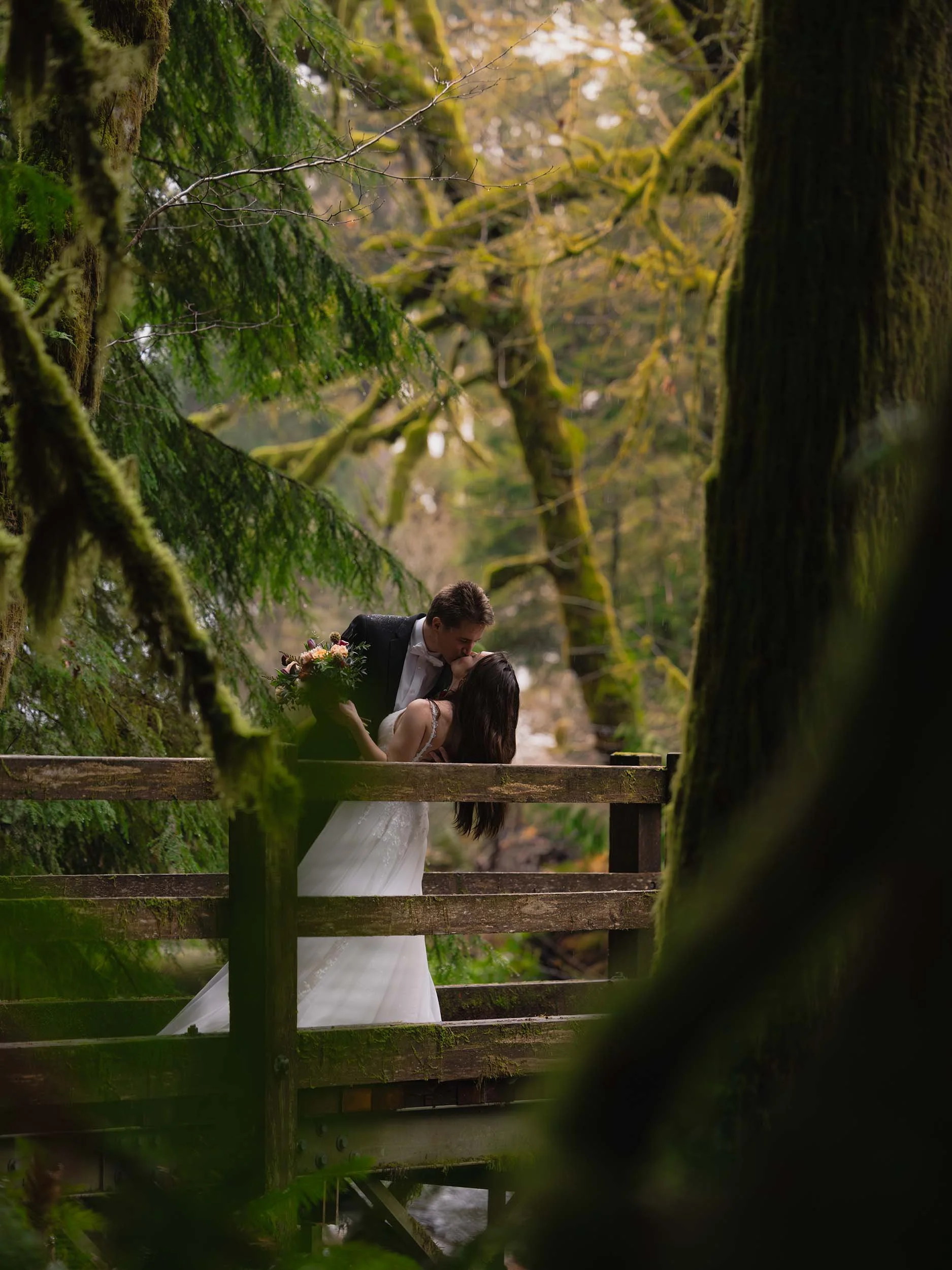 A man and woman in bridal attire kiss on a bridge surrounded by a lush, mossy green forest in Olympic National Park.