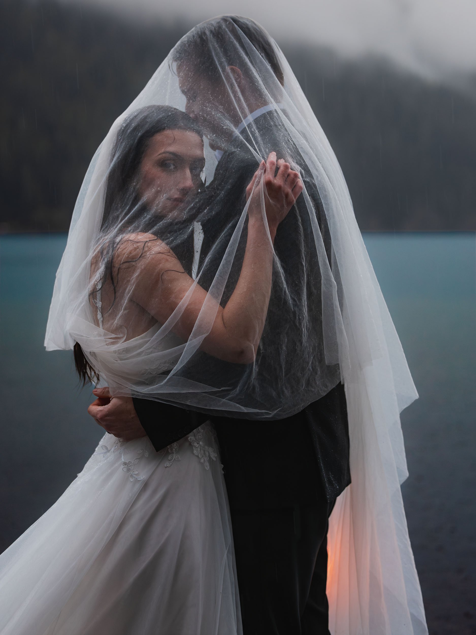 A couple in wedding attire embrace under the bride's wedding veil in the rain during blue hour at Lake Crescent.