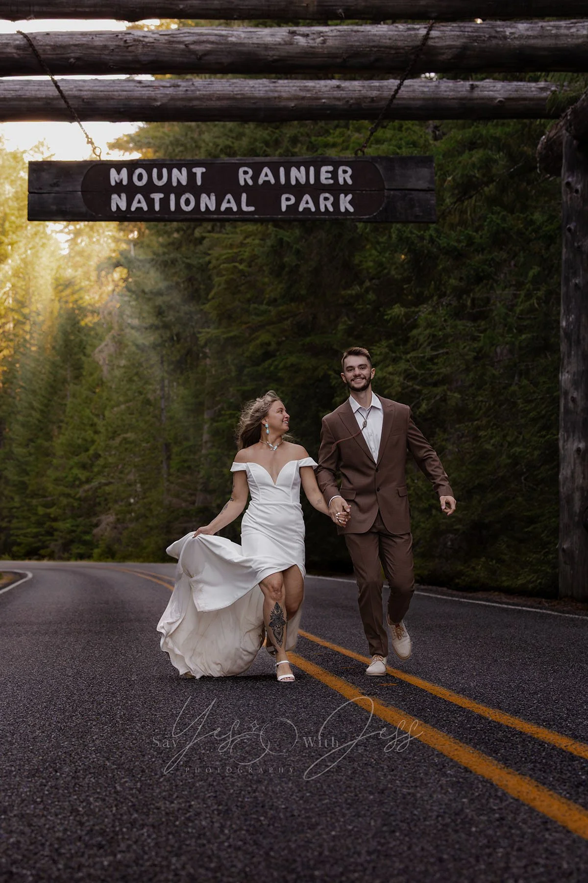 A smiling couple in bridal attire run under the Mount Rainier National Park entrance sign on their wedding day.