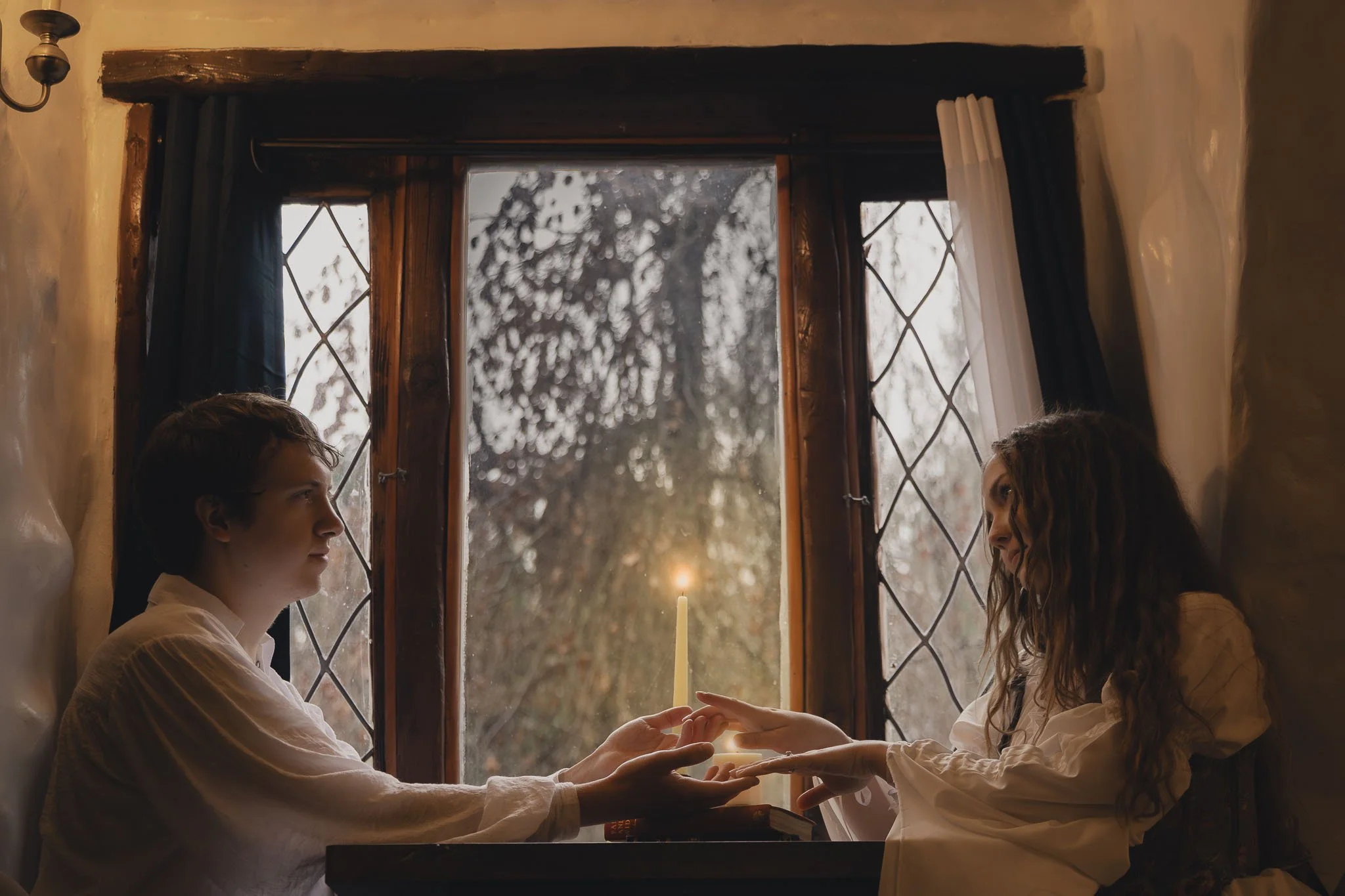 A couple delicately touch hands in front of glowing candlelight, in a room with plaster walls and leaded glass windows during a romance styled session near Olympic National Park.
