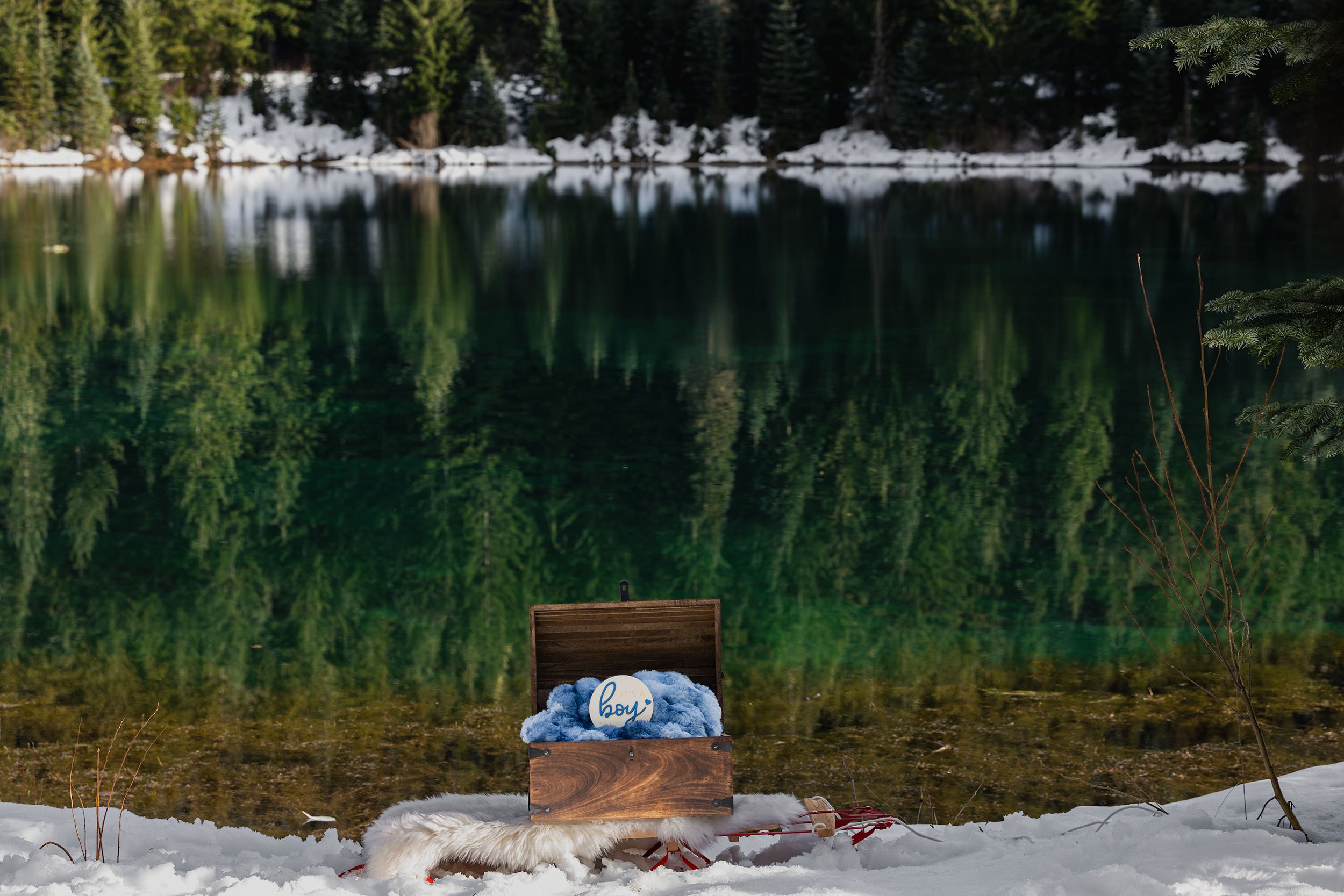 A wood box containing a blue blanket and "boy" sign rests on a sled in the snow at a Gold Creek Pond gender reveal.