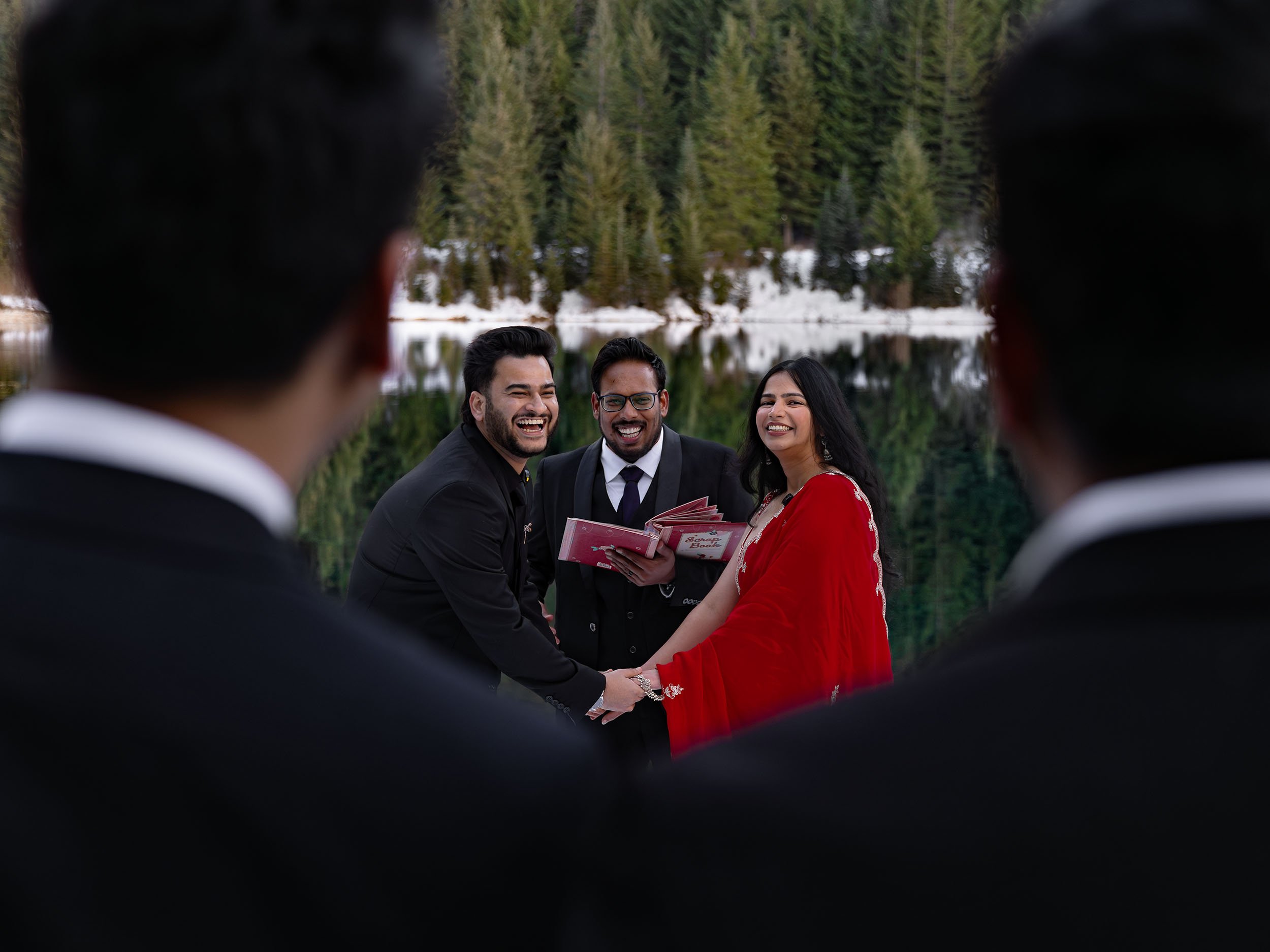 The bride, wearing a red saree, laughs with the groom as seen from a perspective through guests at a Gold Creek Pond micro wedding.