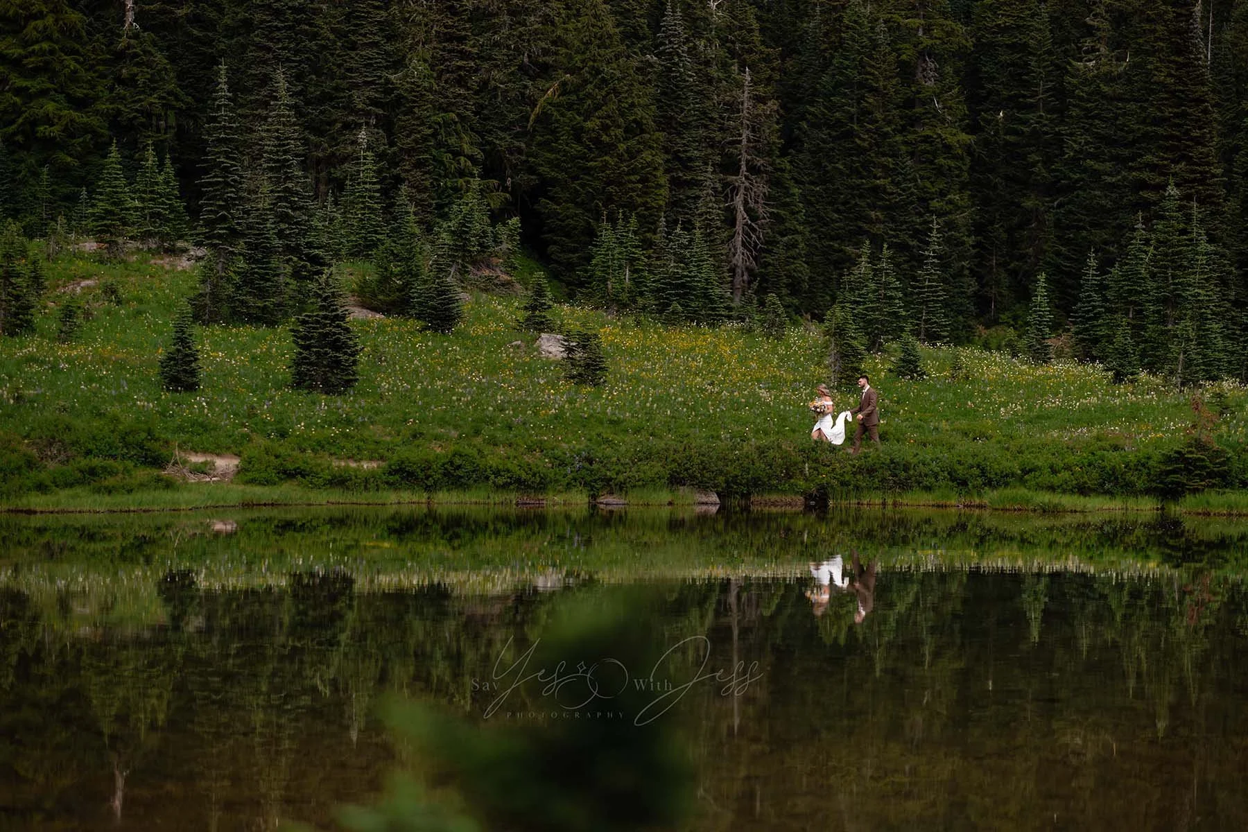 A man holds the train of his bride's wedding dress as they walk around Tipsoo Lake. The scene and the forest and wildflowers are reflected in the lake.