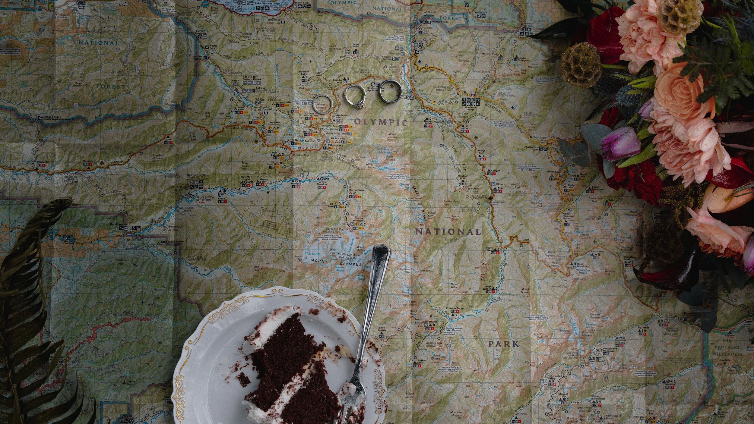 Wedding flat lay consisting of a map of Olympic National Park, wedding rings, wedding cake and a bridal bouquet