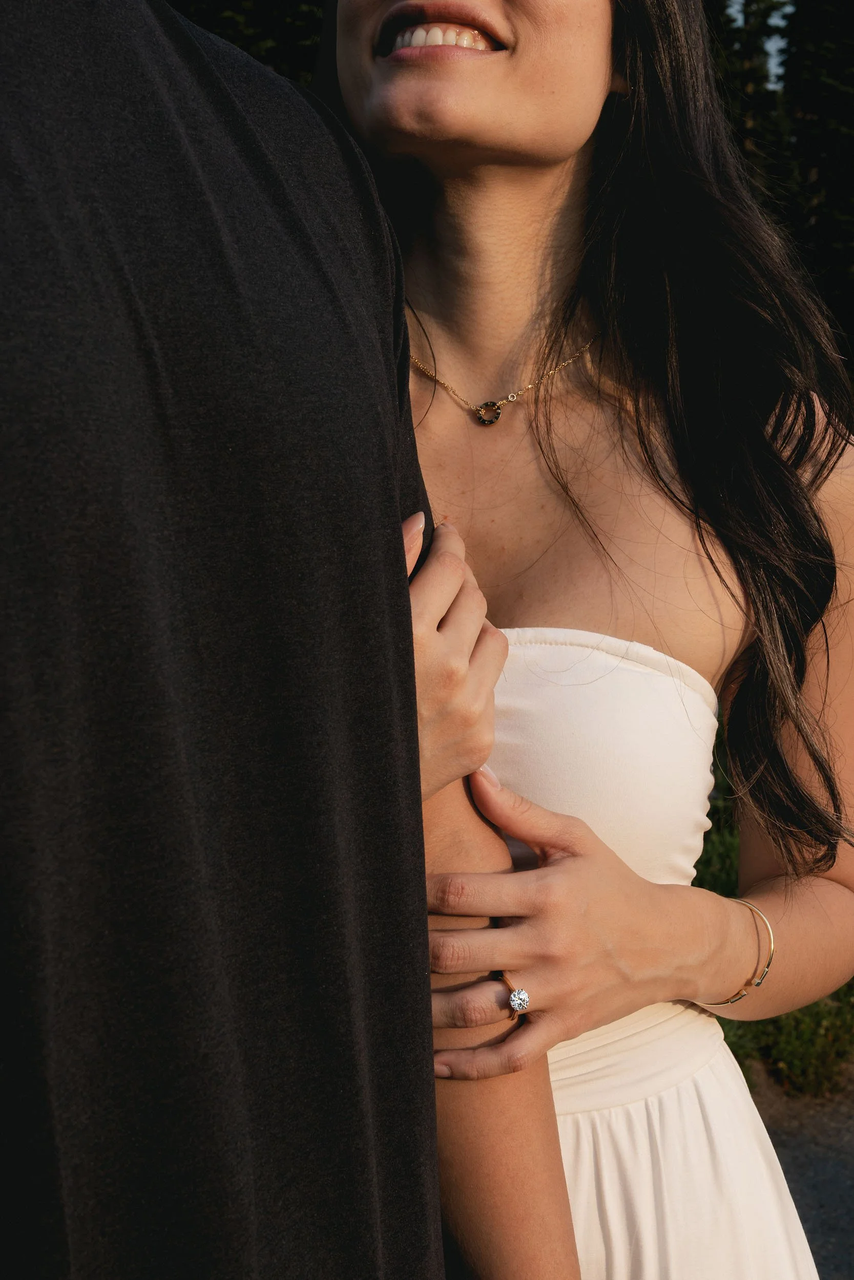 A smiling woman holds onto her fiance's arm, showing her engagement ring at their photography session.