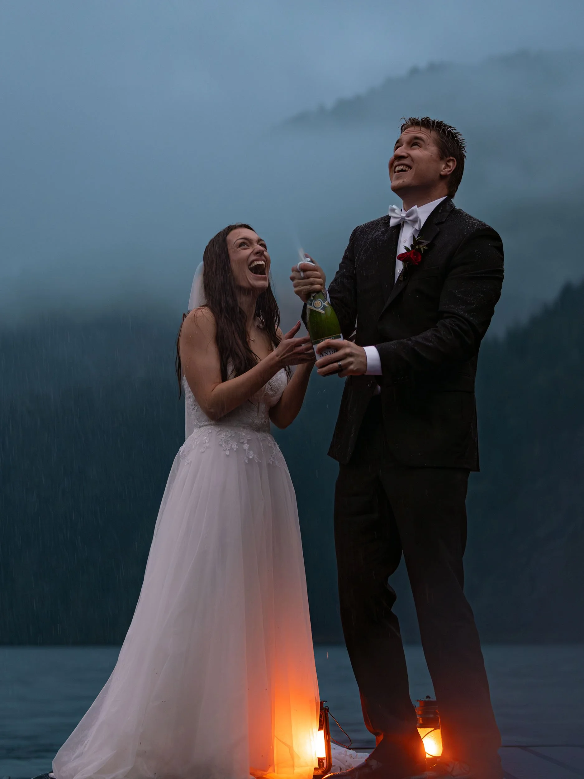 A laughing couple in wedding attire with lanterns spray champagne at blue hour on a dock at Lake Crescent after their wedding. 