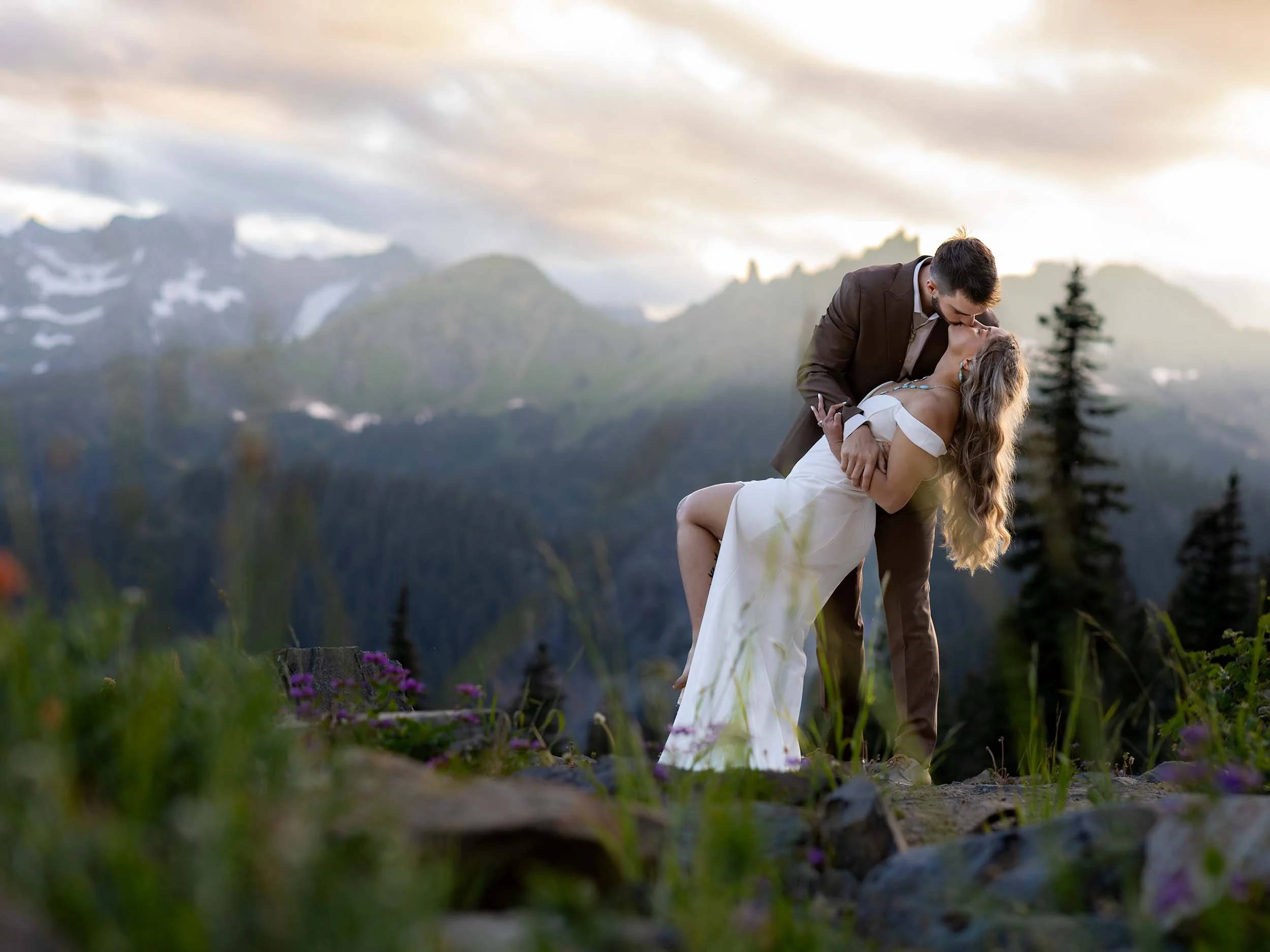 The groom dips his bride at sunset in the cascades surrounded by wildflowers and snow-tipped mountains.