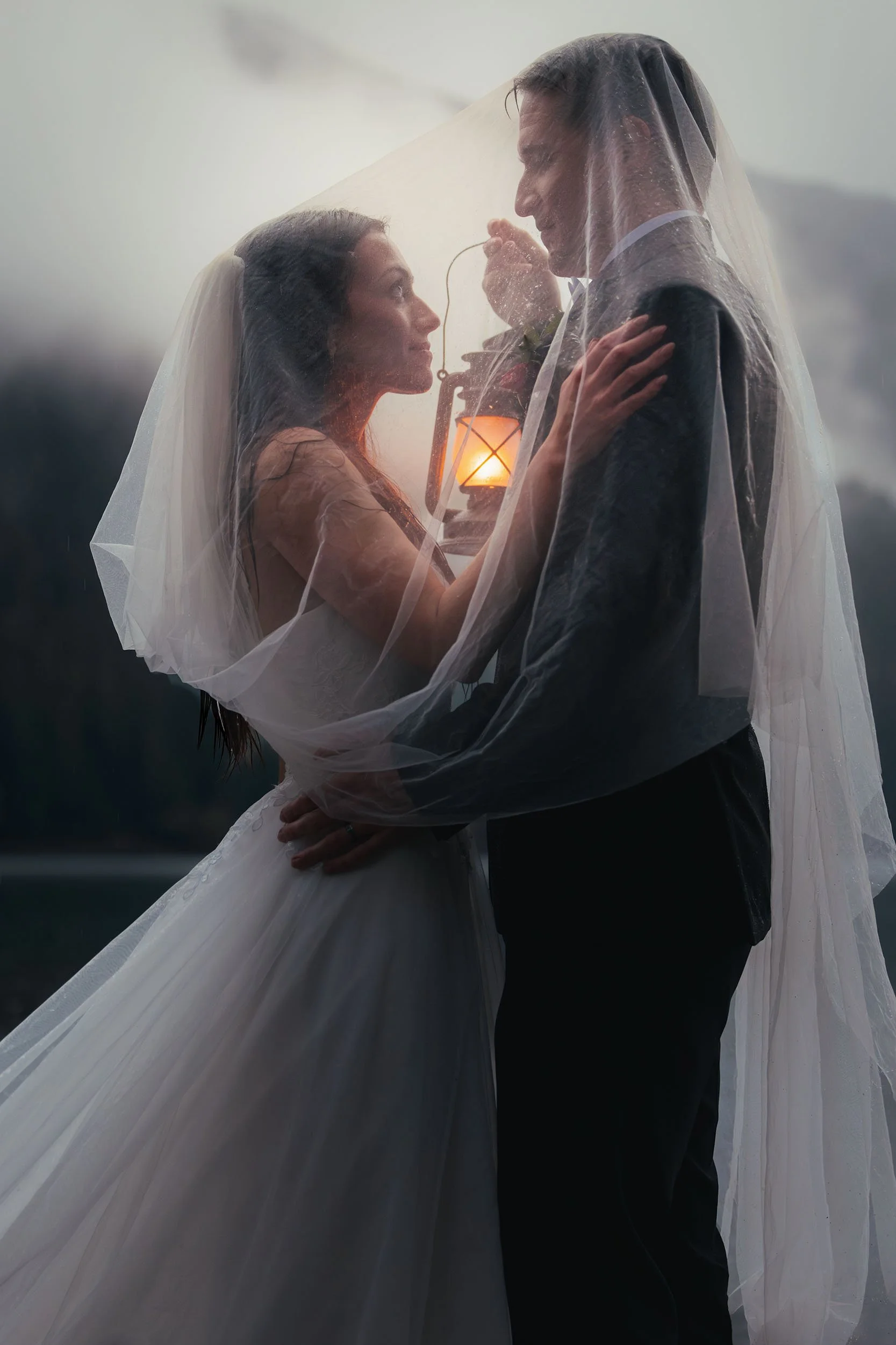 A bride and groom stand under her bridal veil holding a lantern in the rain at Lake Crescent during their elopement.