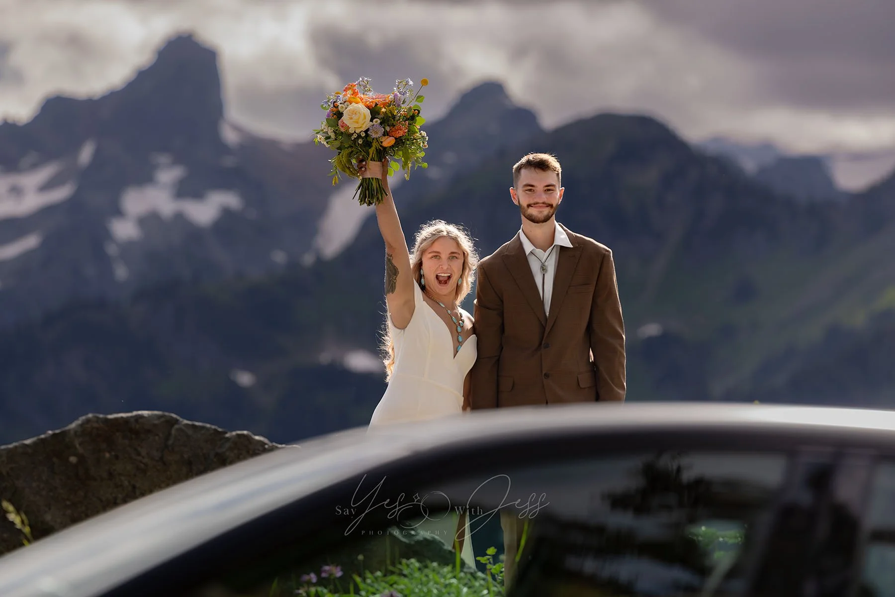 The bride and groom smile at a car riving by honking their horn at their elopement. The bride throws her bouquet arm in the air cheering.
