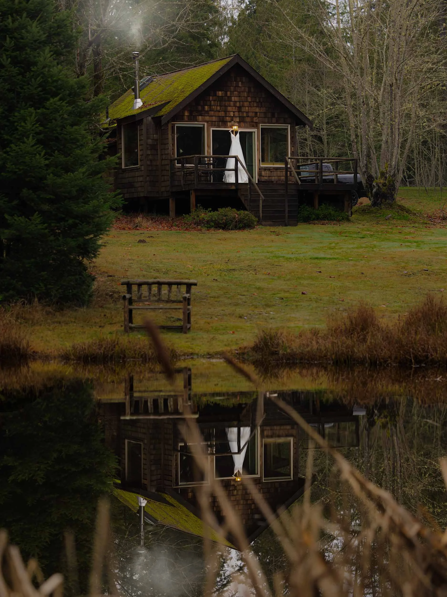 A wedding gown hangs on a cabin porch, as photographed from across a lake perfectly reflecting the moody PNW scene.