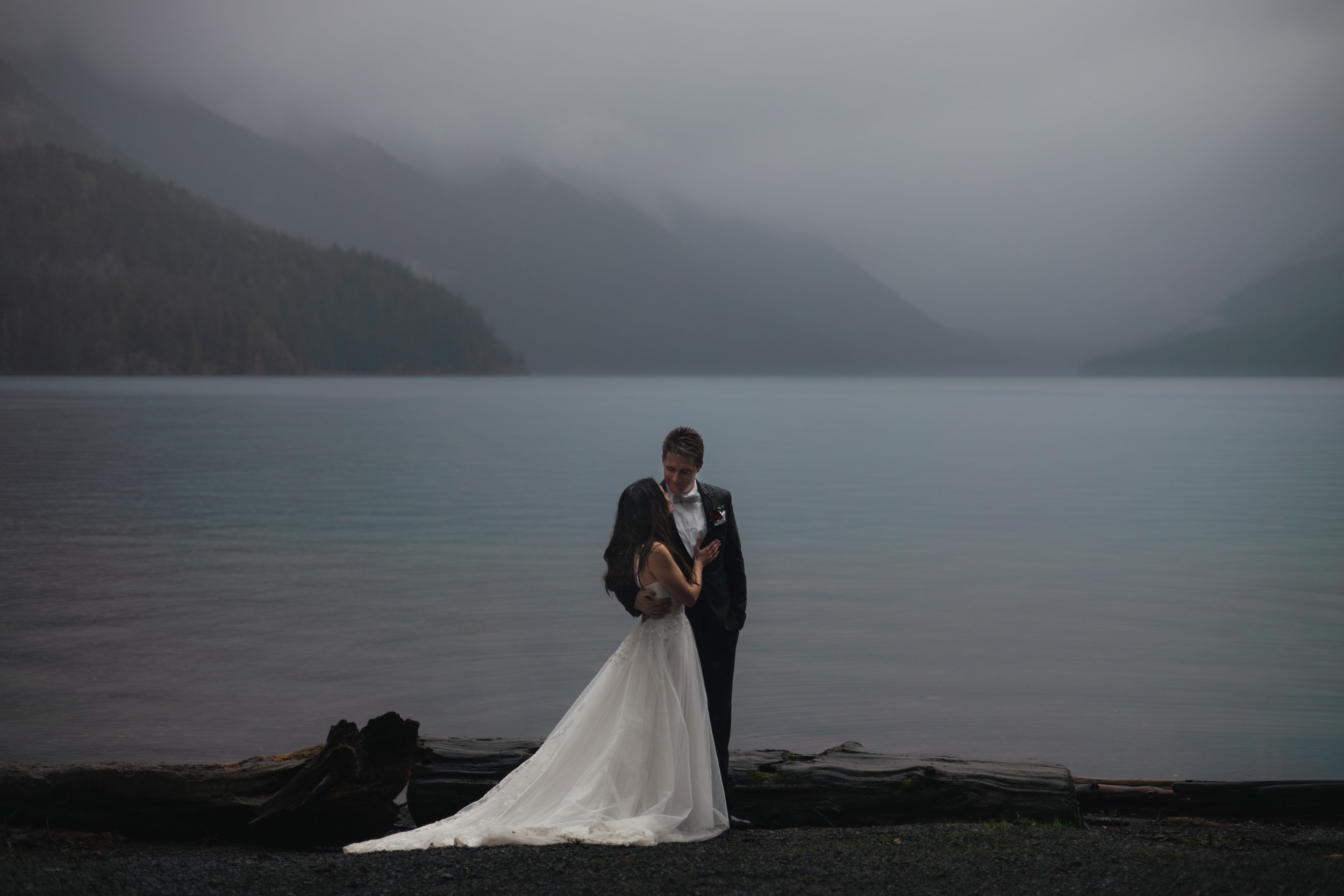 A bride and groom stand in the pouring rain at Lake Crescent during their elopement. The dense fog and blue hour create a dramatic effect.