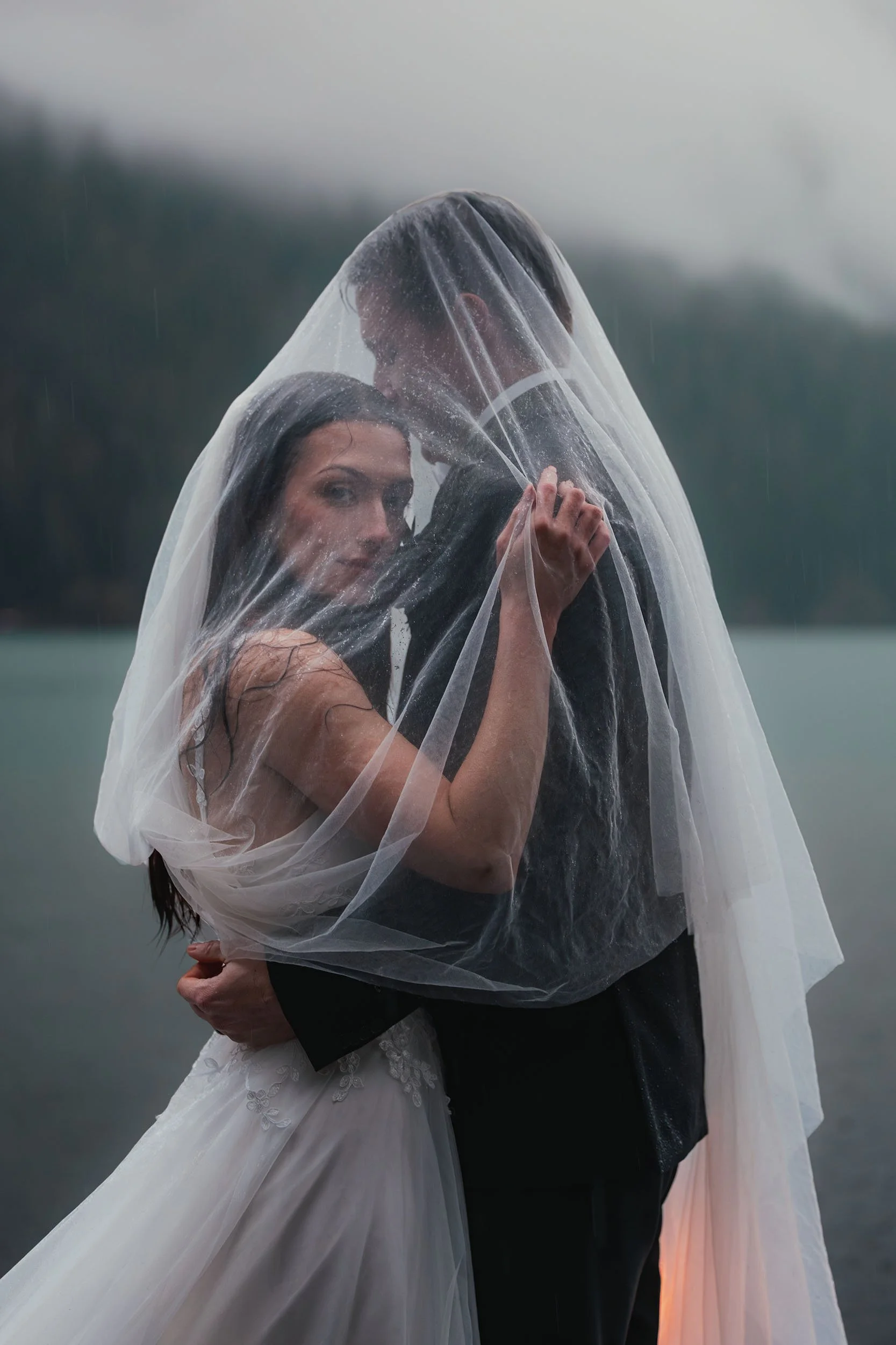 A bride and groom stand under her bridal veil in the rain at Lake Crescent during their elopement. A foggy blue hour enhances the artistic style.
