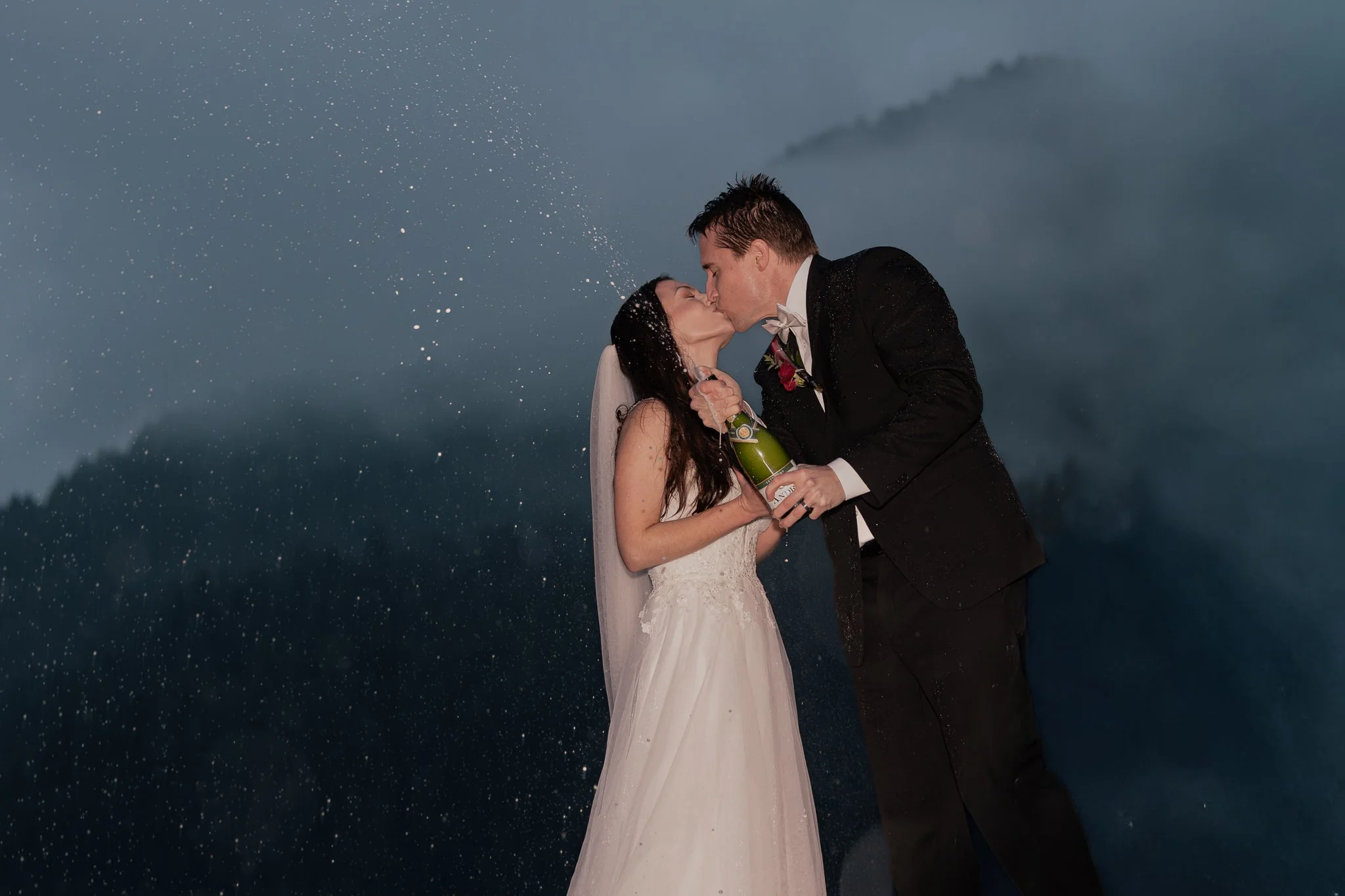 Eloping couple spray champagne and kiss on the dock at Lake Crescent Lodge during a foggy blue hour.