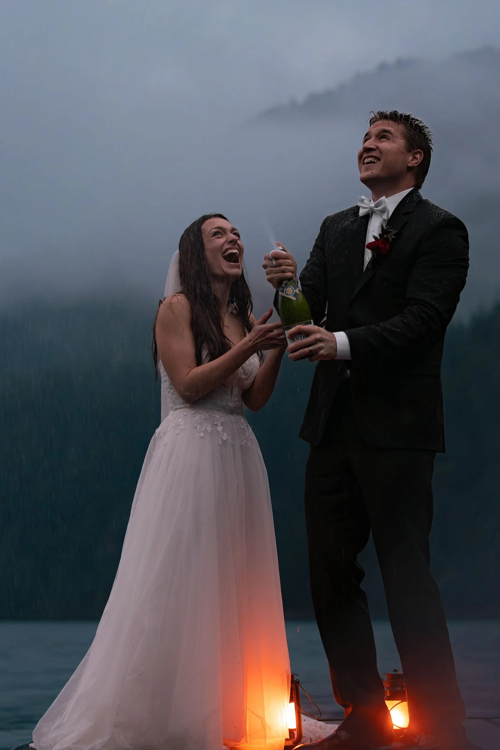 Eloping couple spray champagne on the dock at Lake Crescent during blue hour. They laugh and lanterns glow orange at their feet.