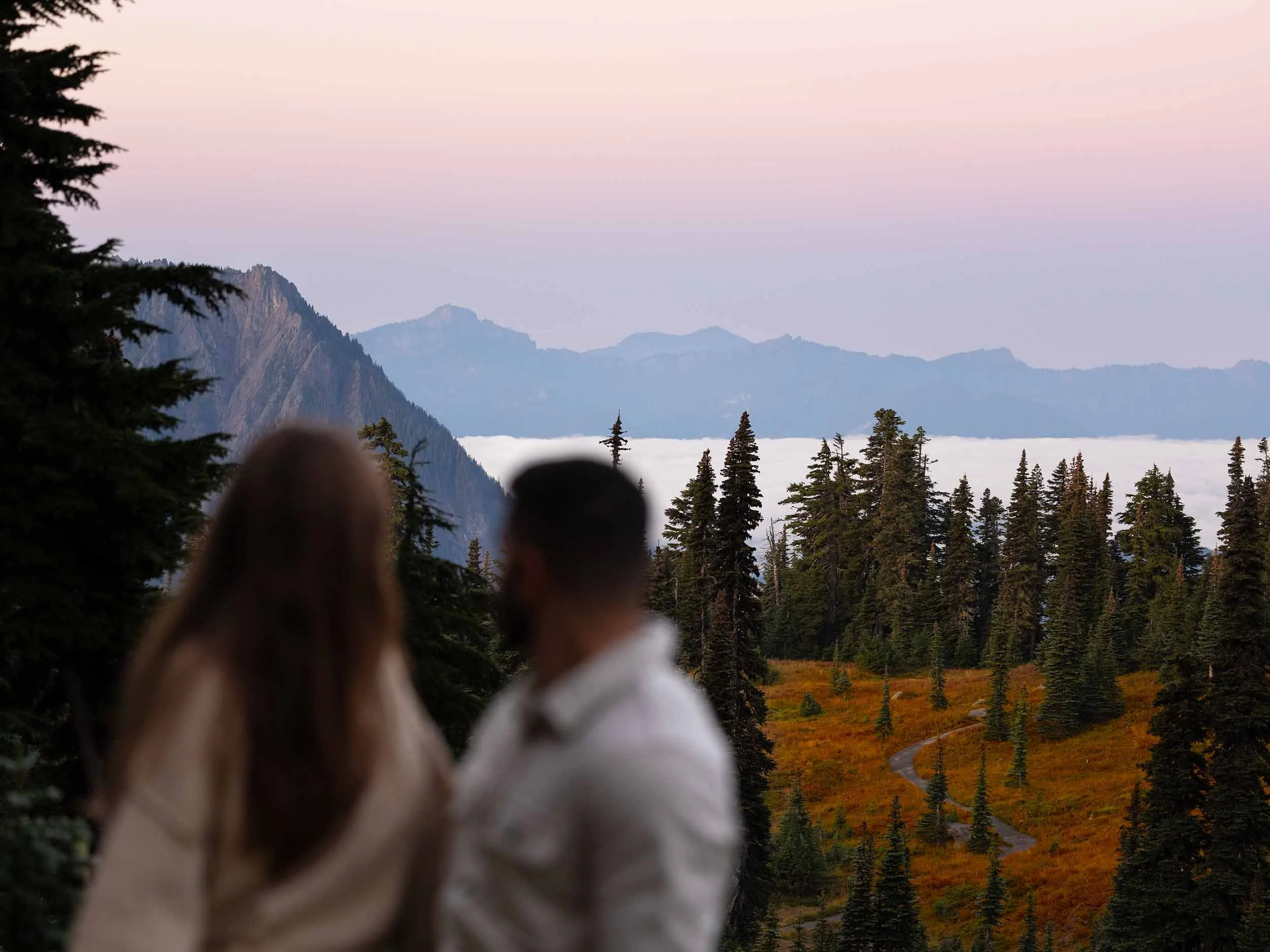 An engaged couple watch a colorful sunrise over the cascade range with a cloud inversion and vibrant fall foliage at Mount Rainier.