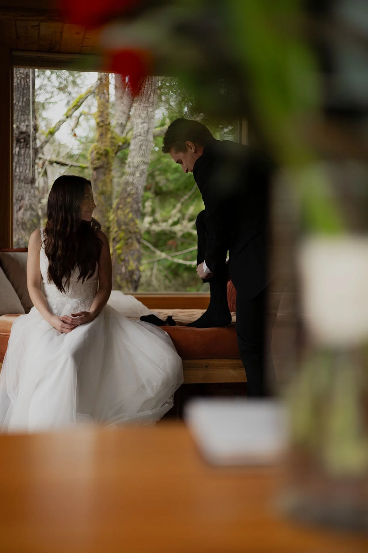 A couple  finish getting ready for their wedding day, from a perspective through a vase with her bridal bouquet in the foreground