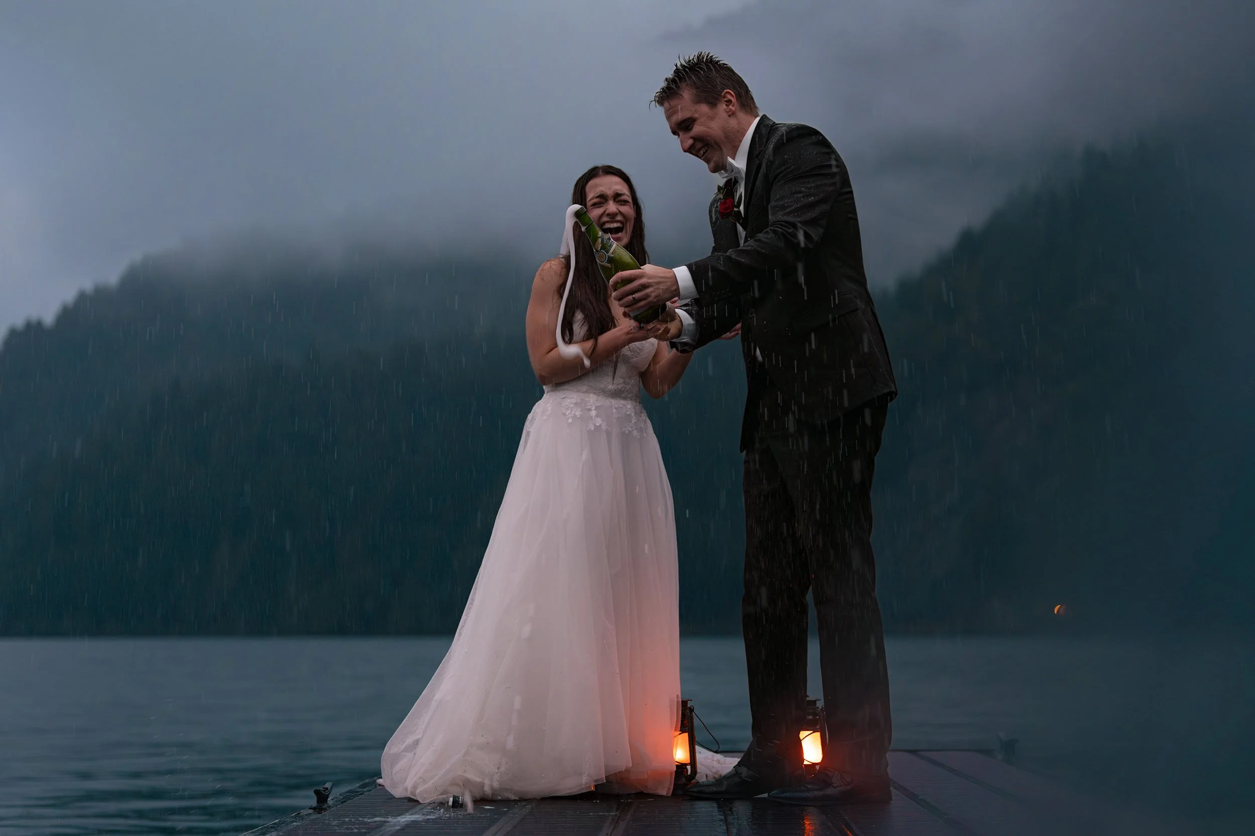 Eloping couple spray champagne on the dock at Lake Crescent during blue hour. They laugh and lanterns glow orange at their feet.
