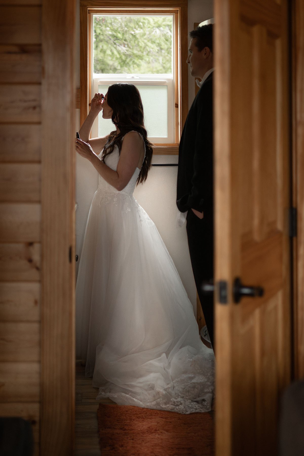 A couple in wedding attire prepare for their elopement in a Port Angeles cabin. The bride applies mascara in the bathroom mirror.