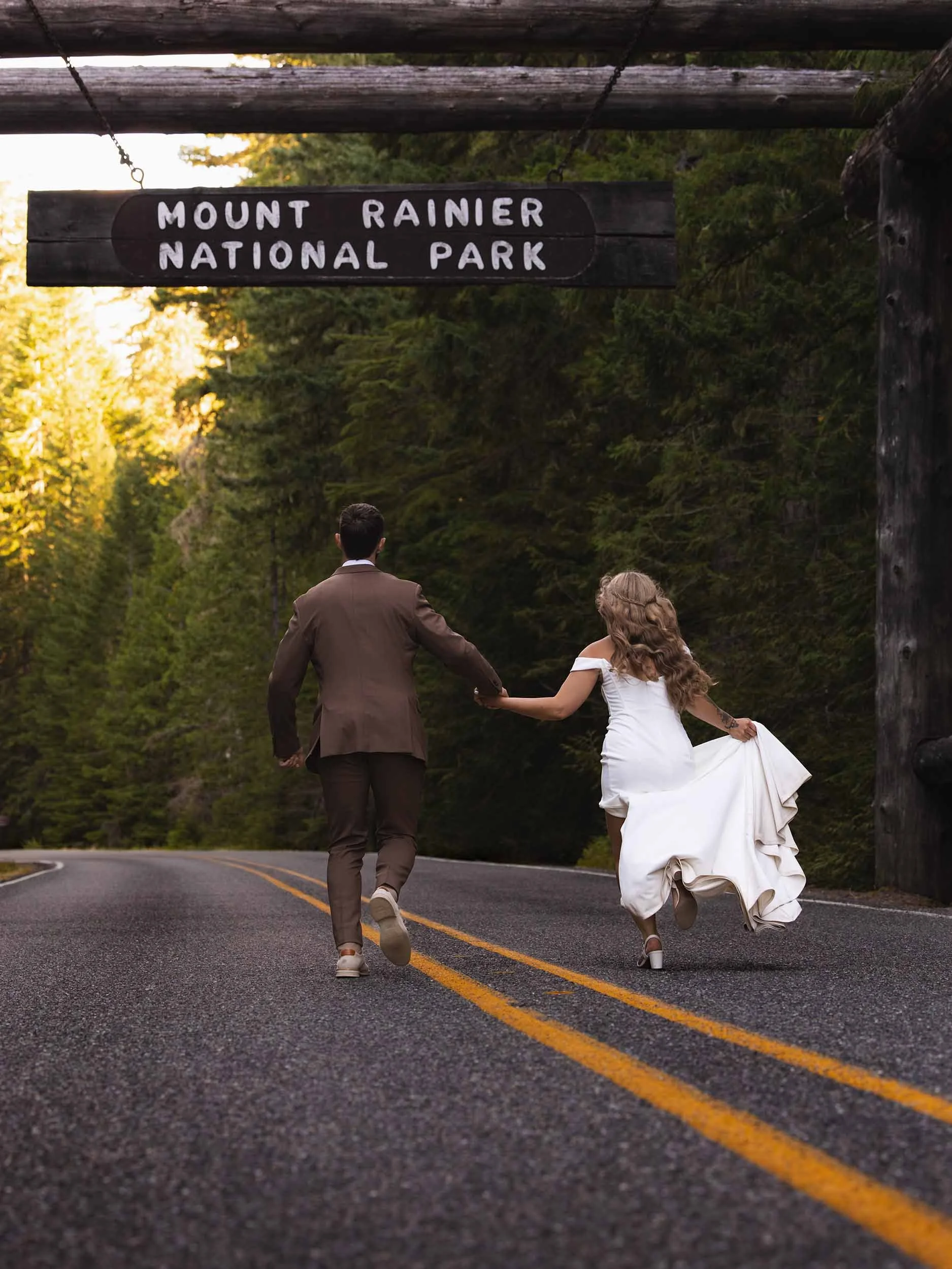 A wedding couple run under the Mount Rainier National Park entrance sign during their summer elopement.