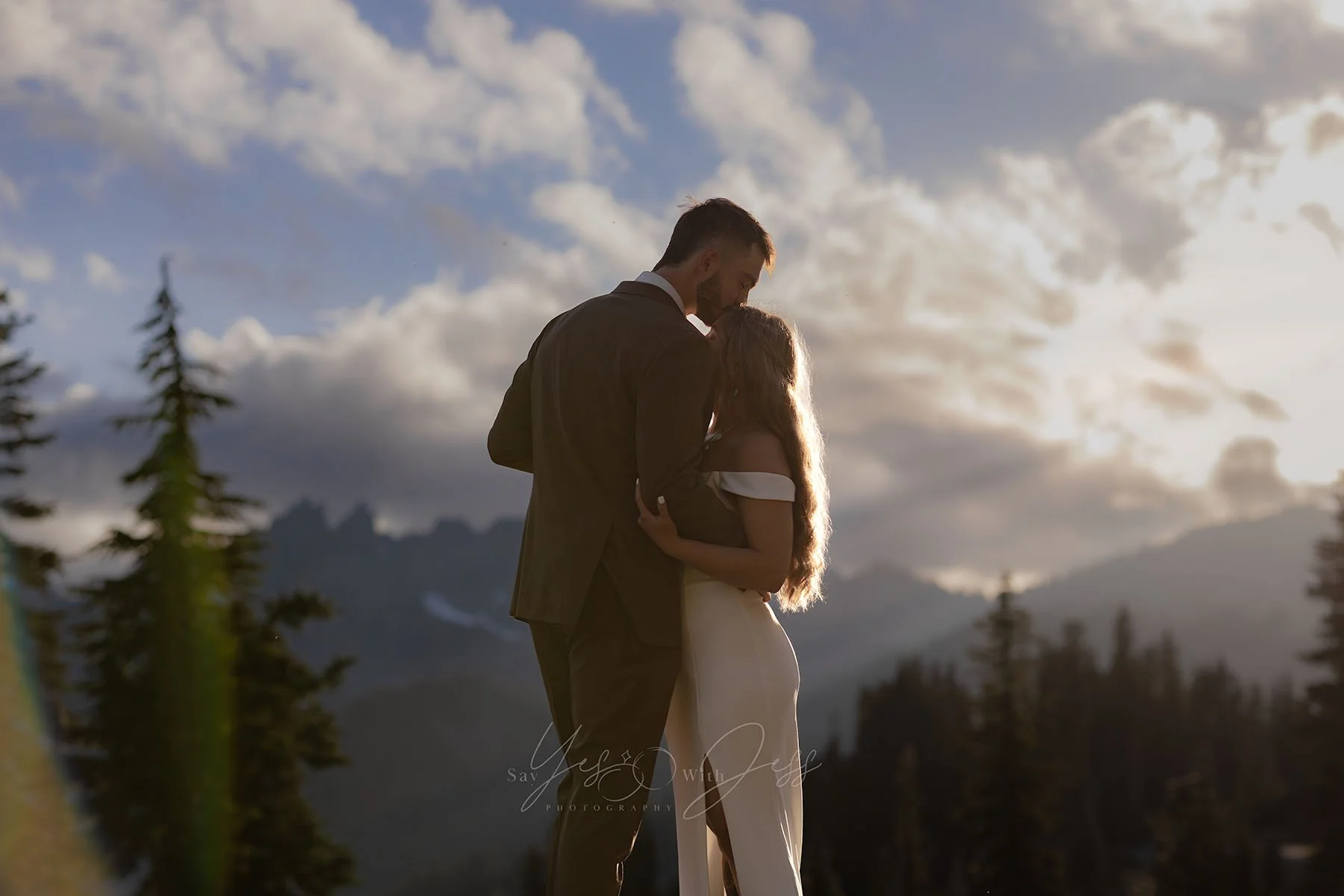 A bride and groom are backlit by the sun and mountains as the man kisses his bride's forehead on their elopement day at Mount Rainier.
