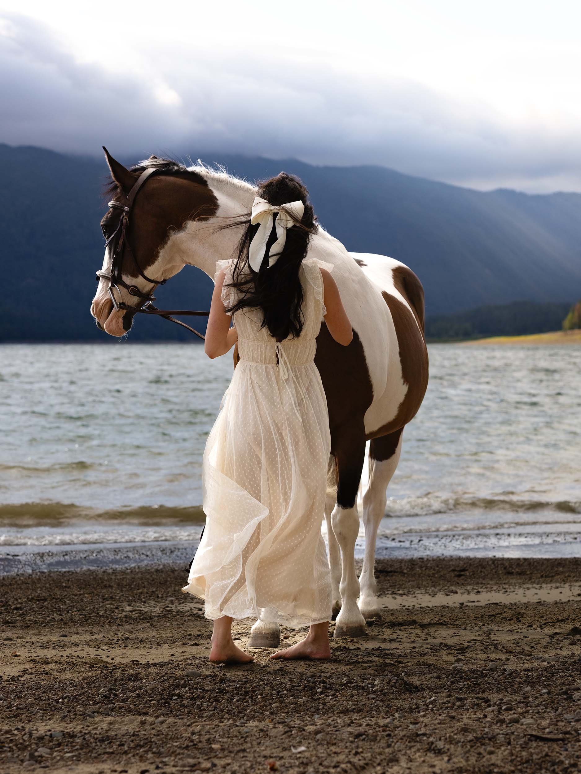 A woman wearing a flowy ivory dress and bow in her hair stands with a horse at Speelyi Beach for a creative editorial photoshoot