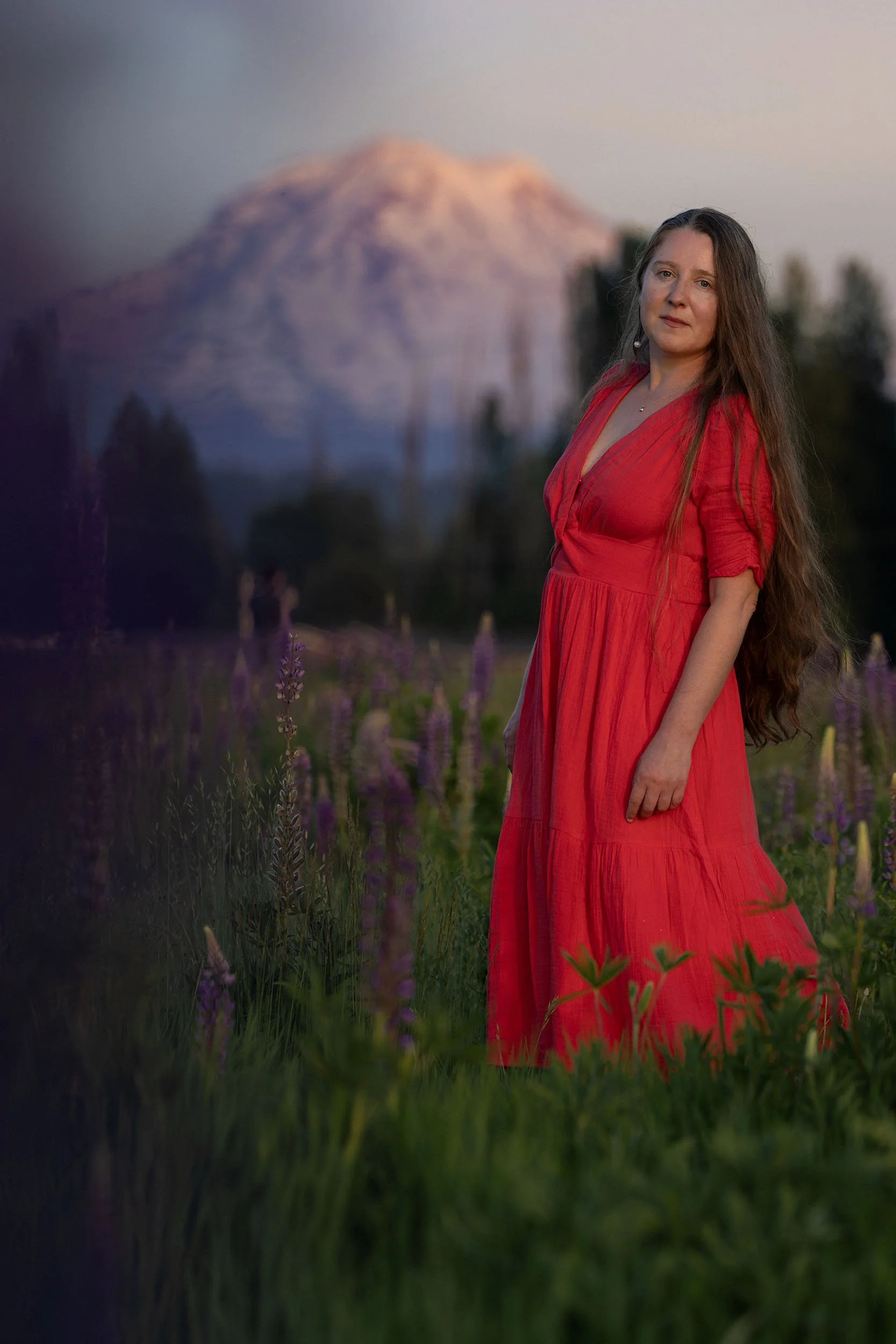 A woman in a pink dress stands in a field of purple lupine at sunset. Mount Rainier looms large in the background.