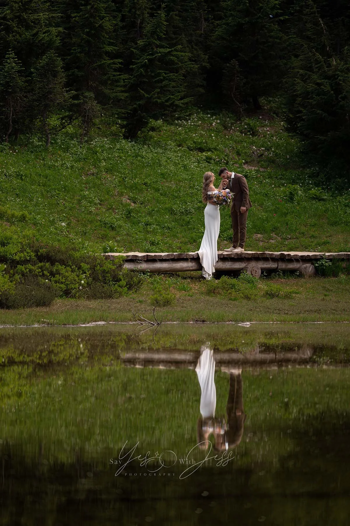 A man kisses his bride's hand. They stand on a wood bridge, framed by the scenes reflection in a pond at Mount Rainier.