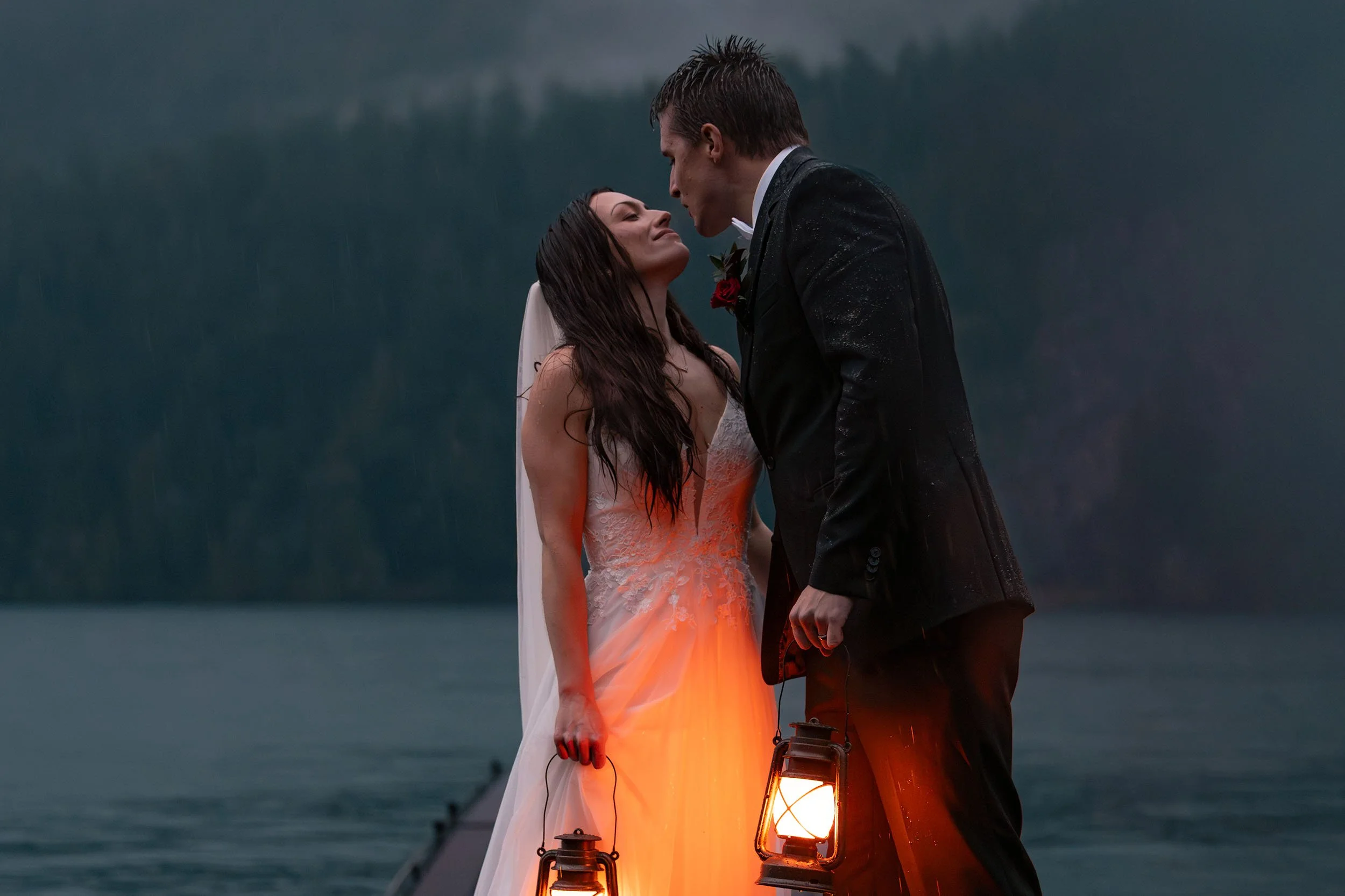Eloping stand in the rain on the dock at Lake Crescent during blue hour. They smile at each other and hold lanterns glowing orange.