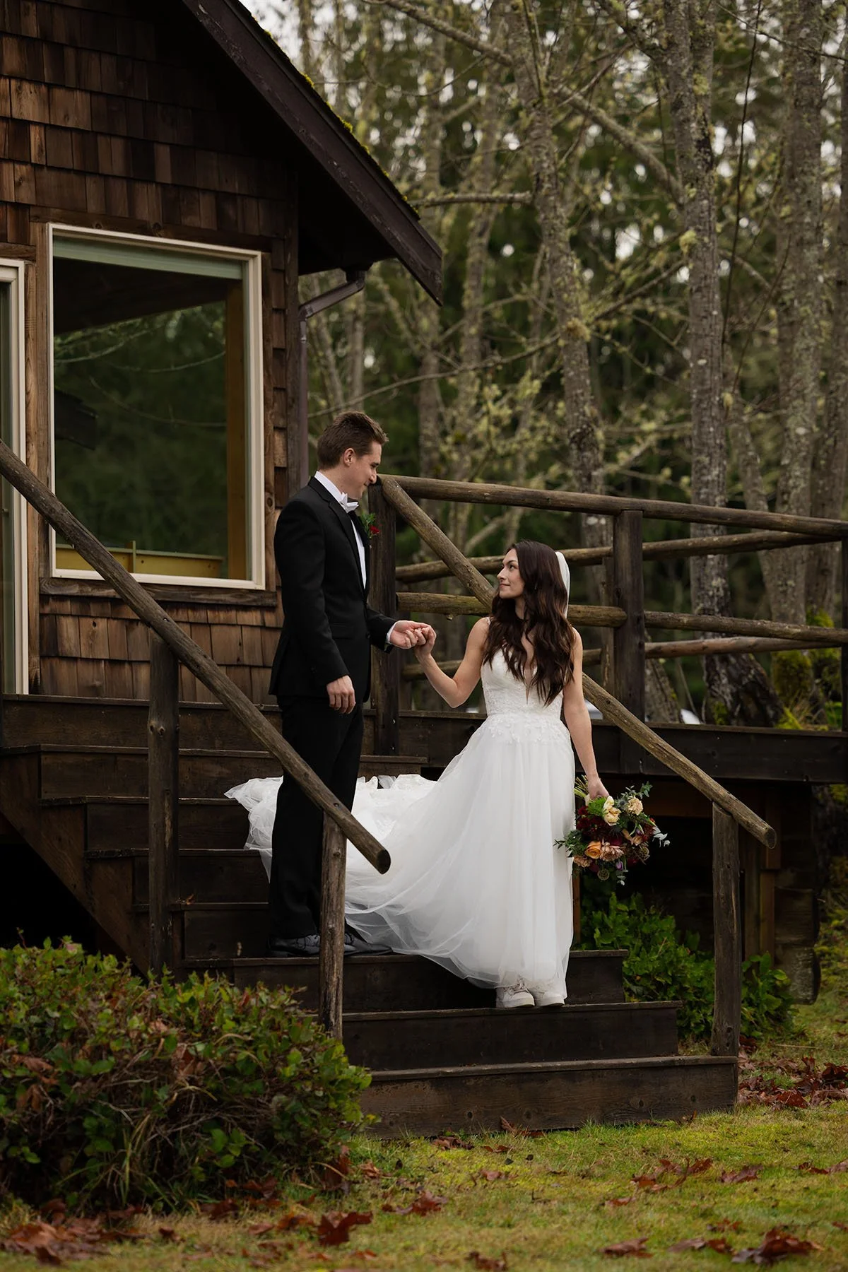 A couple in wedding attire hold hands and walk down the steps of a Port Angeles cabin