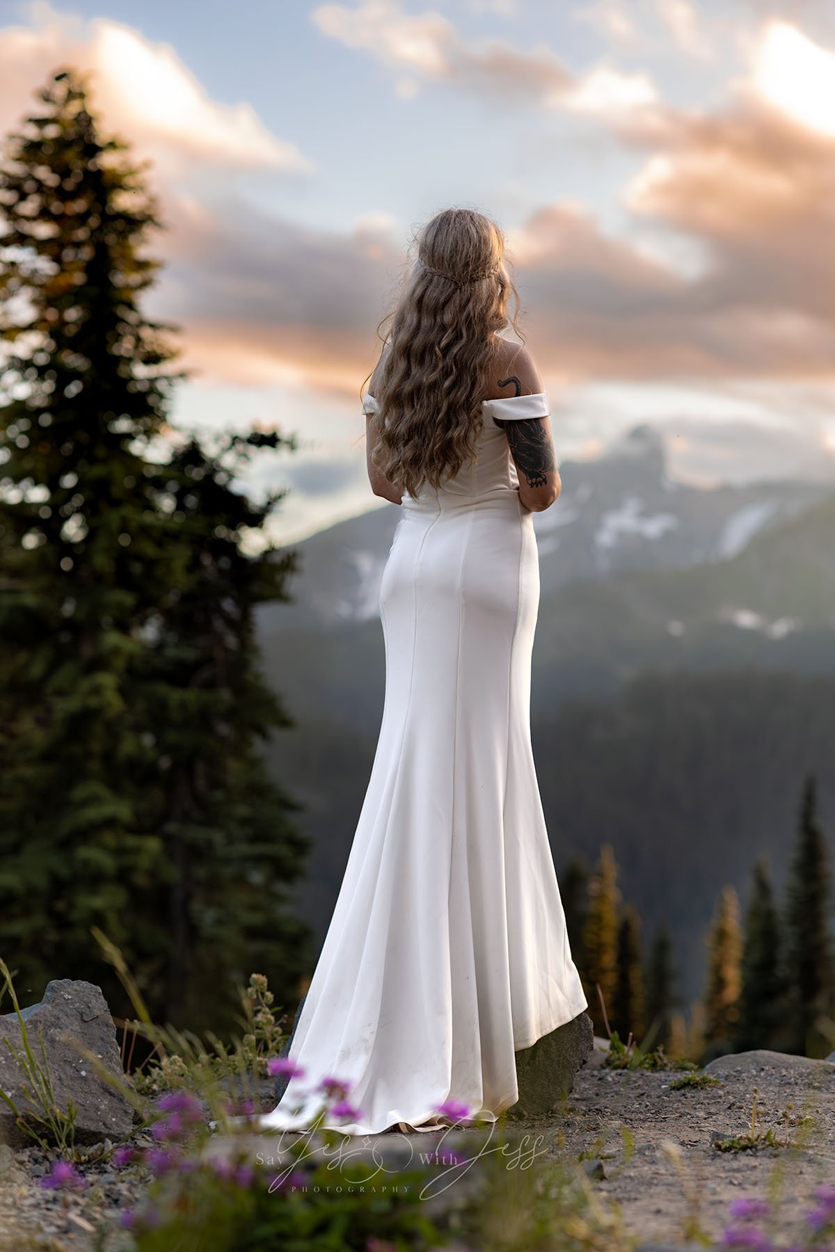 A bride with long, wavy blonde hair stands on a rock at sunset looking at Mount Rainier on her wedding day.