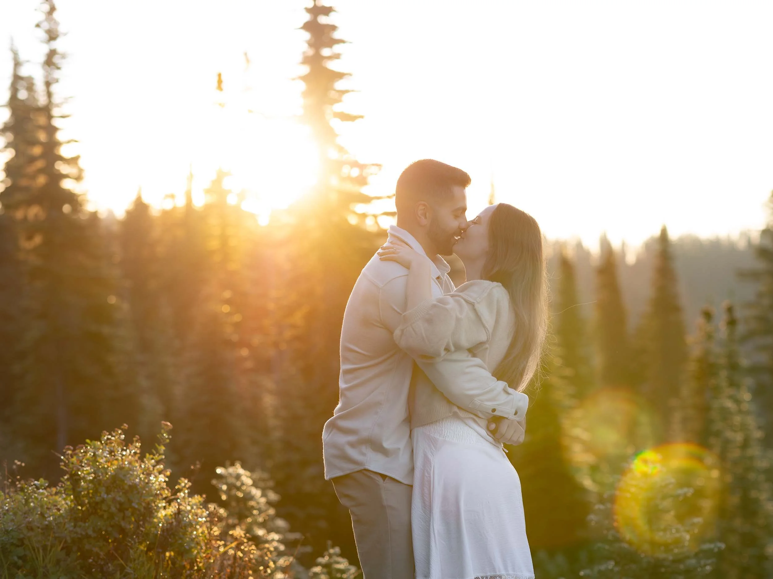The sine rises over the treeline at Mount Rainier , creating an orange glow on a kissing couple at an engagement photography session.