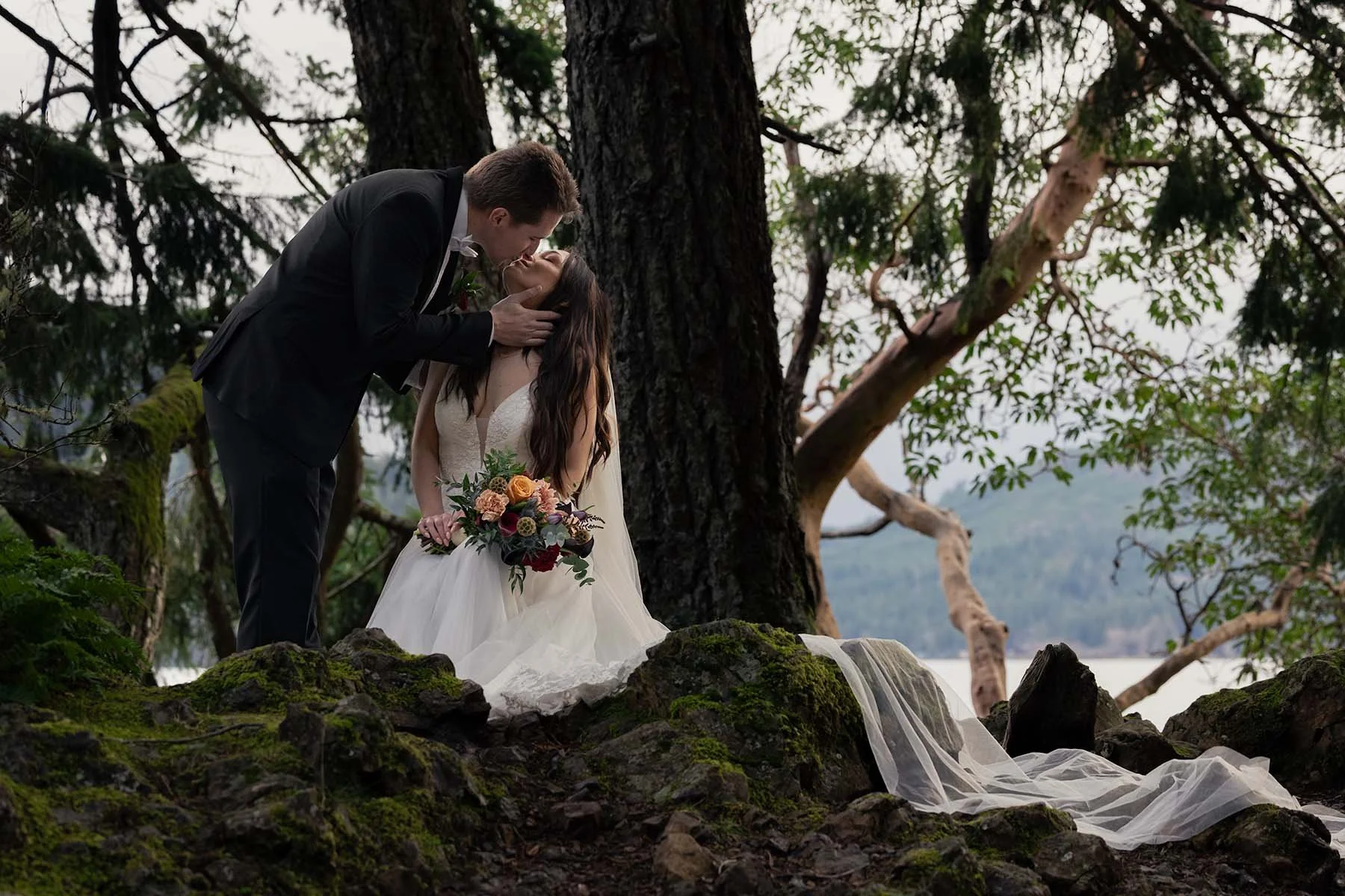 A couple in wedding attire kiss. The bride sits on mossy rocks and her veil flows over them with Lake Crescent in the background