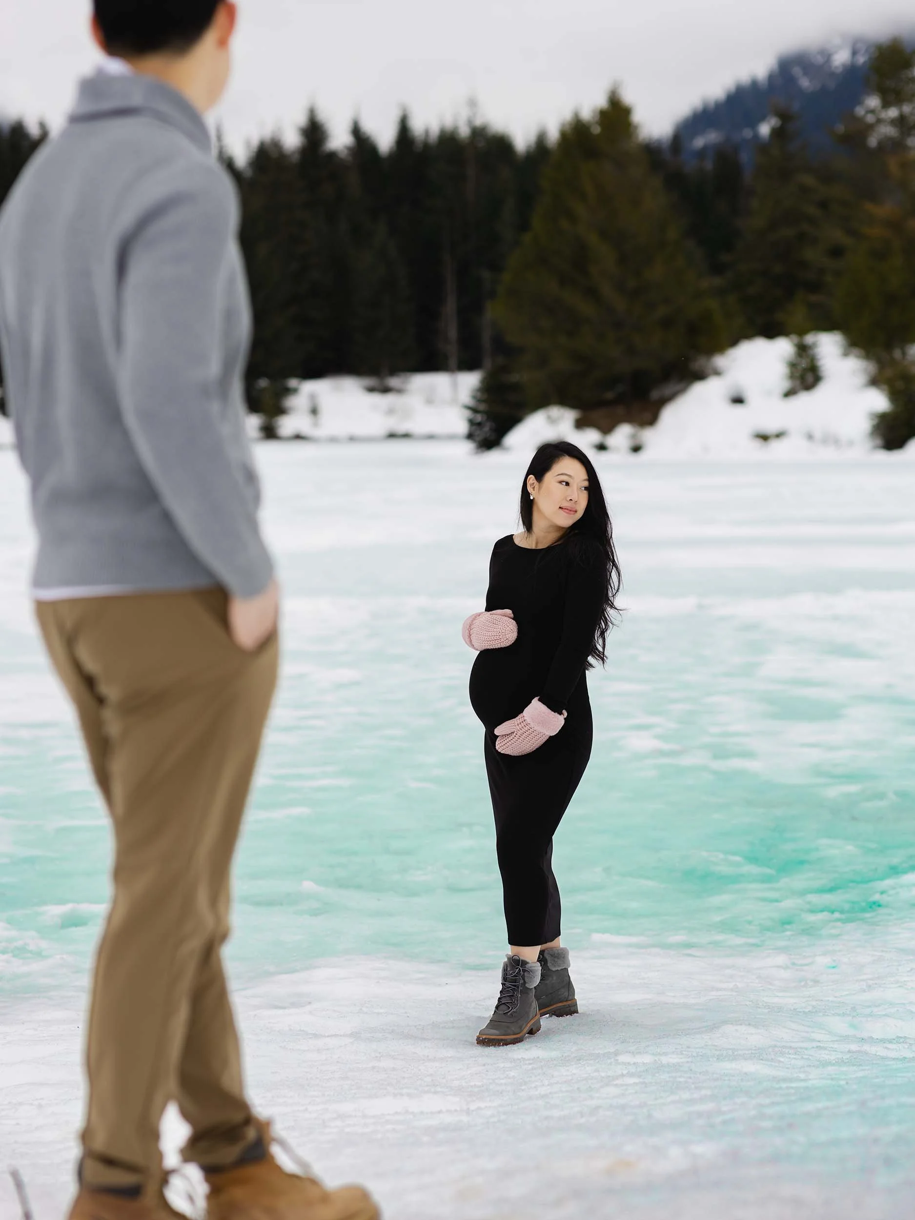 Maternity couple stand at the edge of a frozen, turquoise Gold Creek Pond. Mountains are blanketed in a moody PNW fog. 