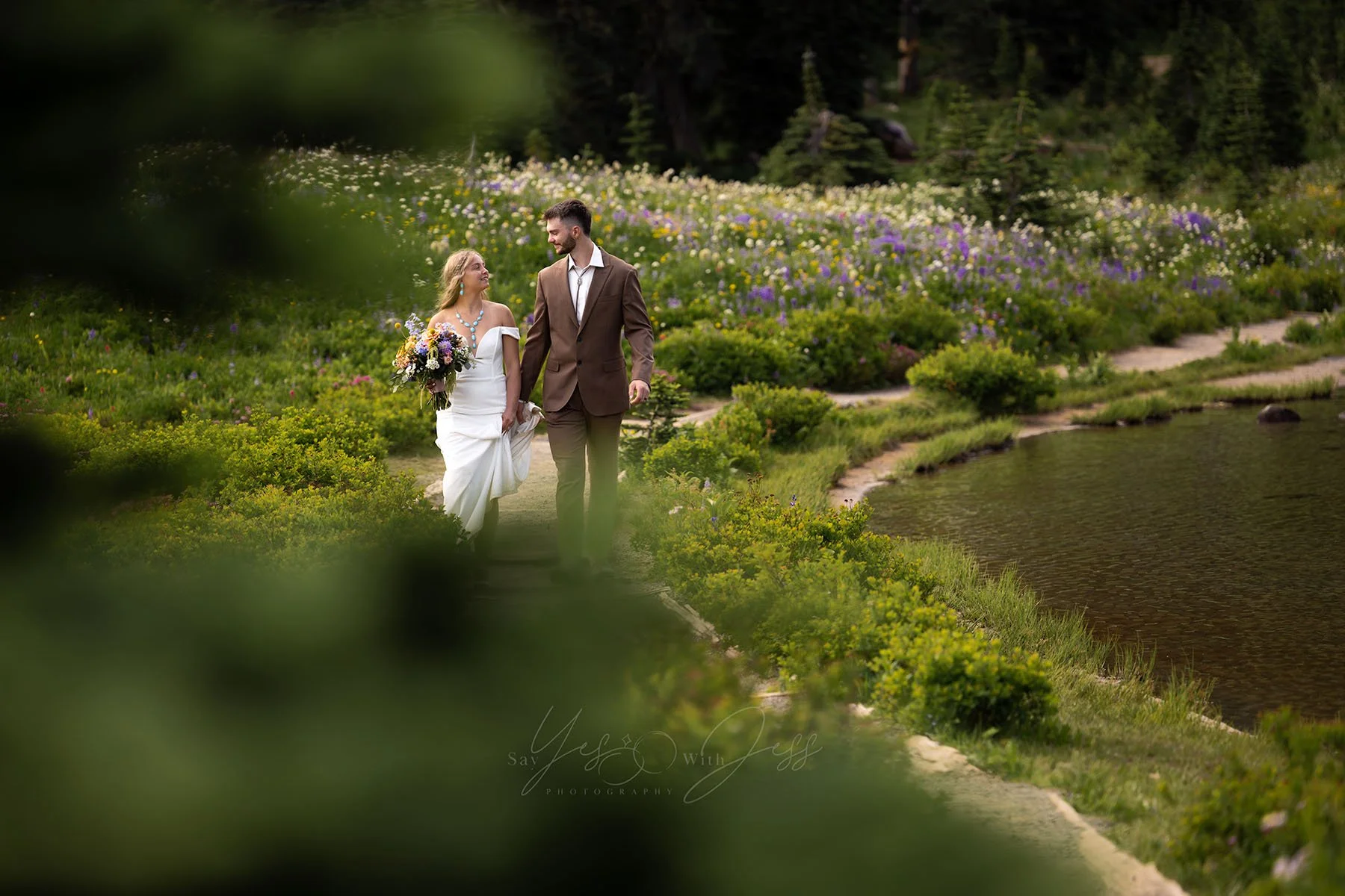 Bride and groom hold hands walking around Tipsoo Lake on their elopement day. They are framed by green pine trees in the foreground and surrounded by vibrant subalpine wildflowers.
