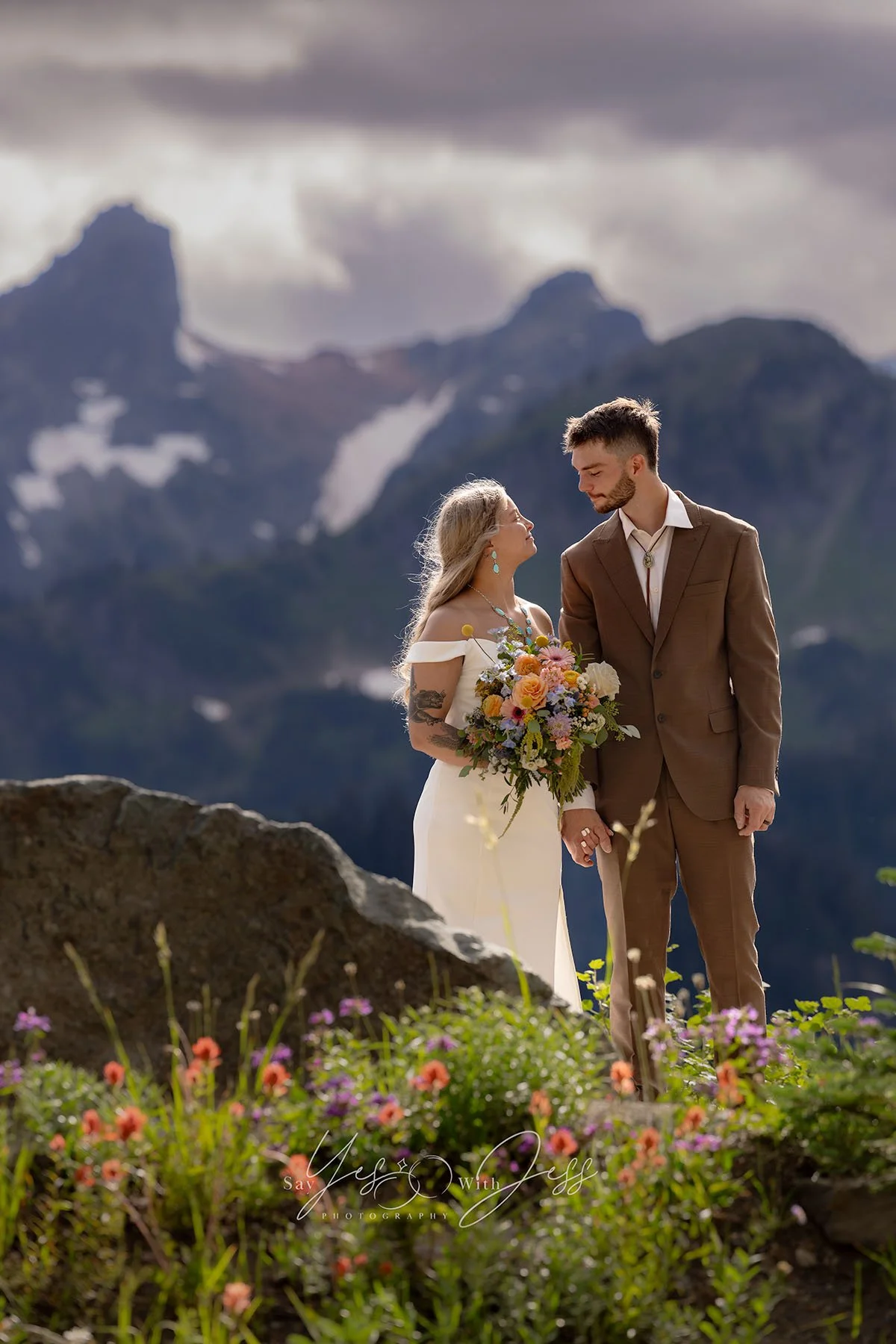 A bride and groom are surrounded by colorful wildflowers and mountains on their elopement day. They smile at each other and hold hands.
