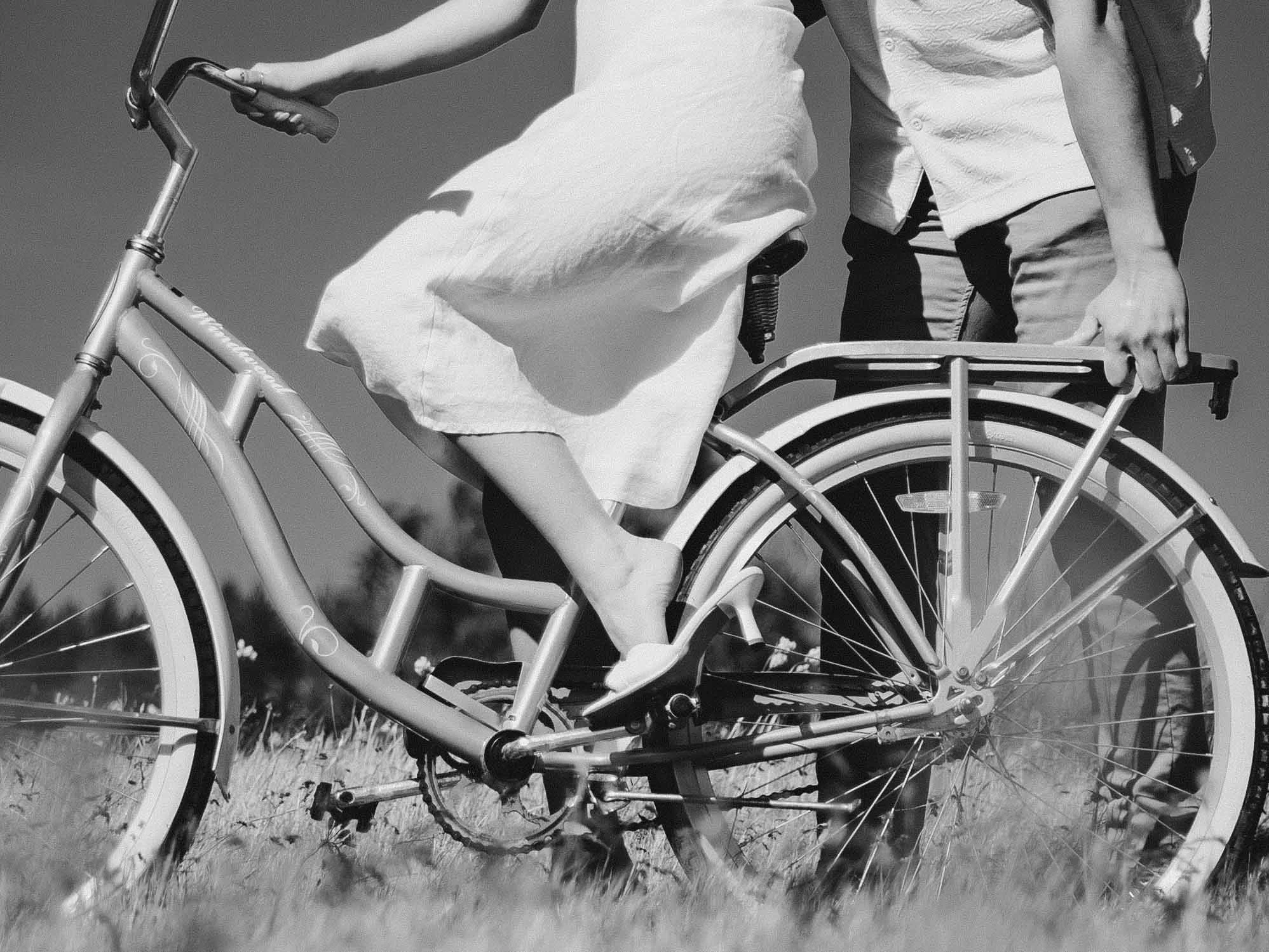 Retro black and white crop of a girl in a white dress and heels sitting on a bike as her fiance holds it steady.