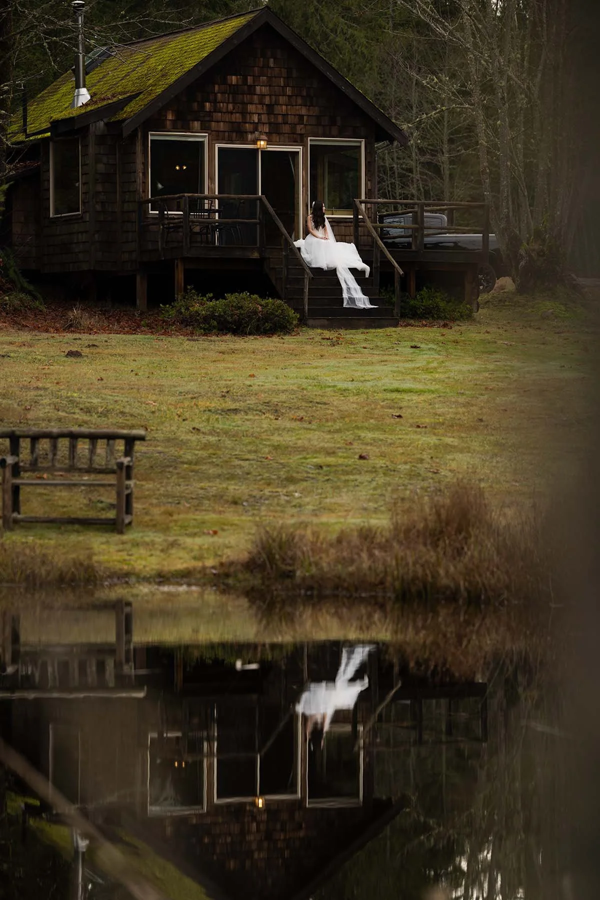 A bride in a wedding gown sits on the steps of a cabin. The scene is reflected in a pond on the property.