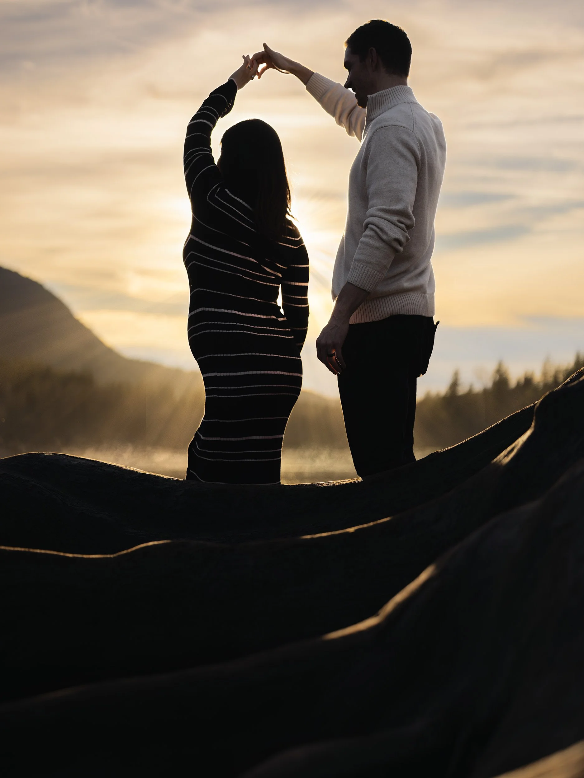 Maternity couple dance between old growth tree roots at Rattlesnake Lake. The setting sun rays glow orange and backlight the couple.
