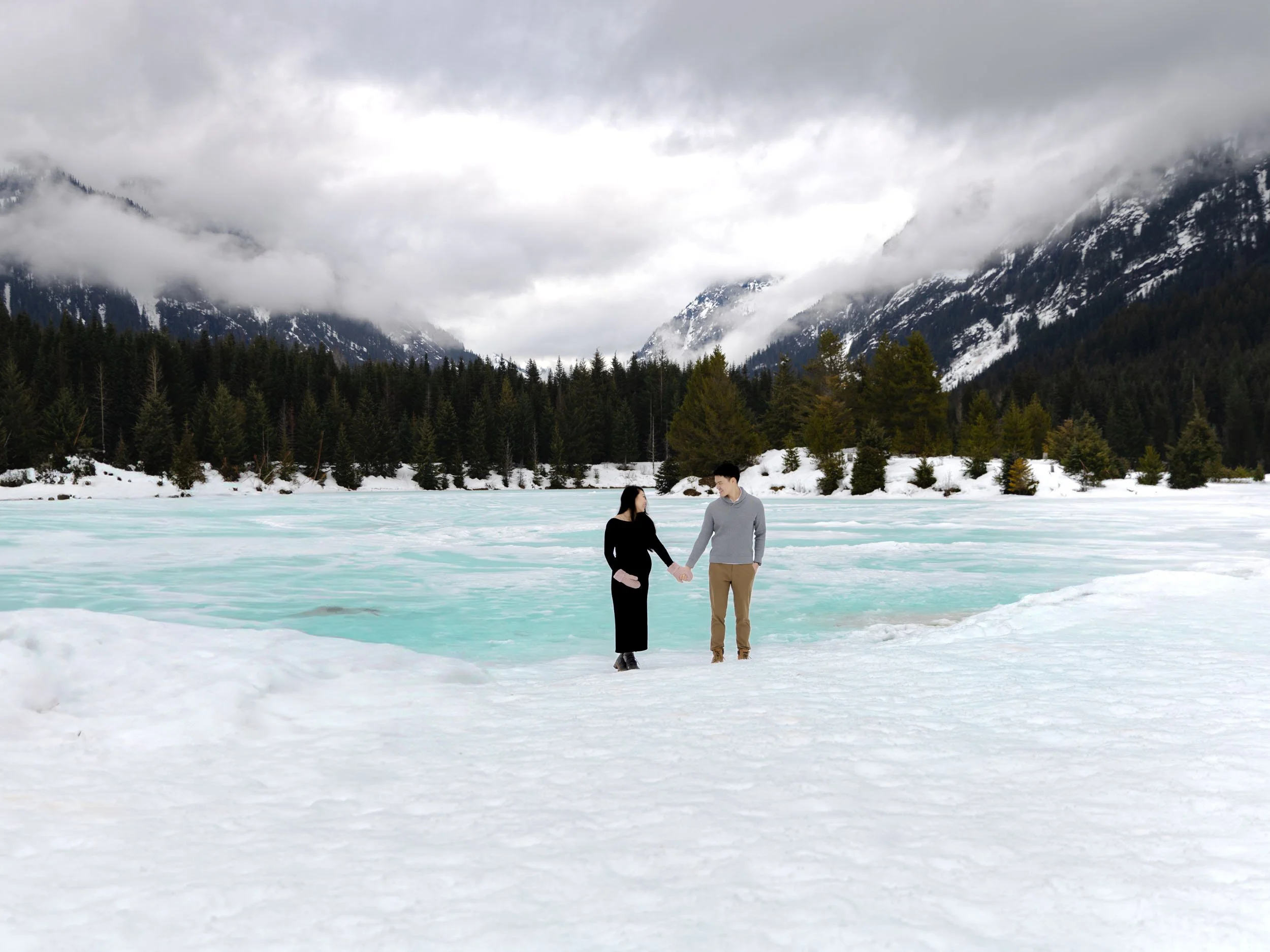 Amy and Jerald hold hands in the snow in front of a frozen, turquoise Gold Creek Pond. The mountains are blanketed with moody PNW fog.
