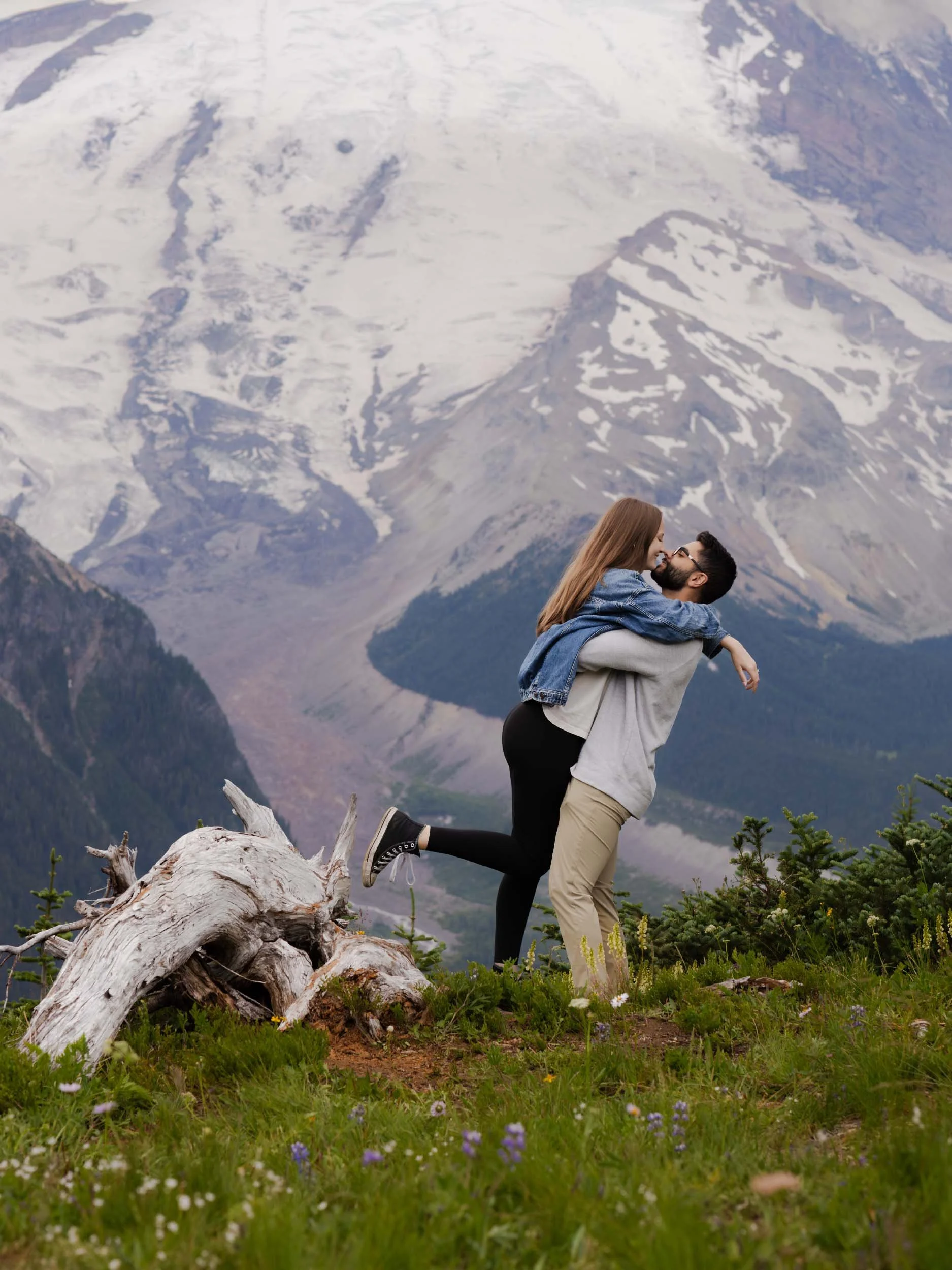A couple in casual clothes embracing outdoors in a mountainous area with snow-capped peaks and green grass with wildflowers. James lifts his fiance in front of Mount Rainier after proposing marriage.