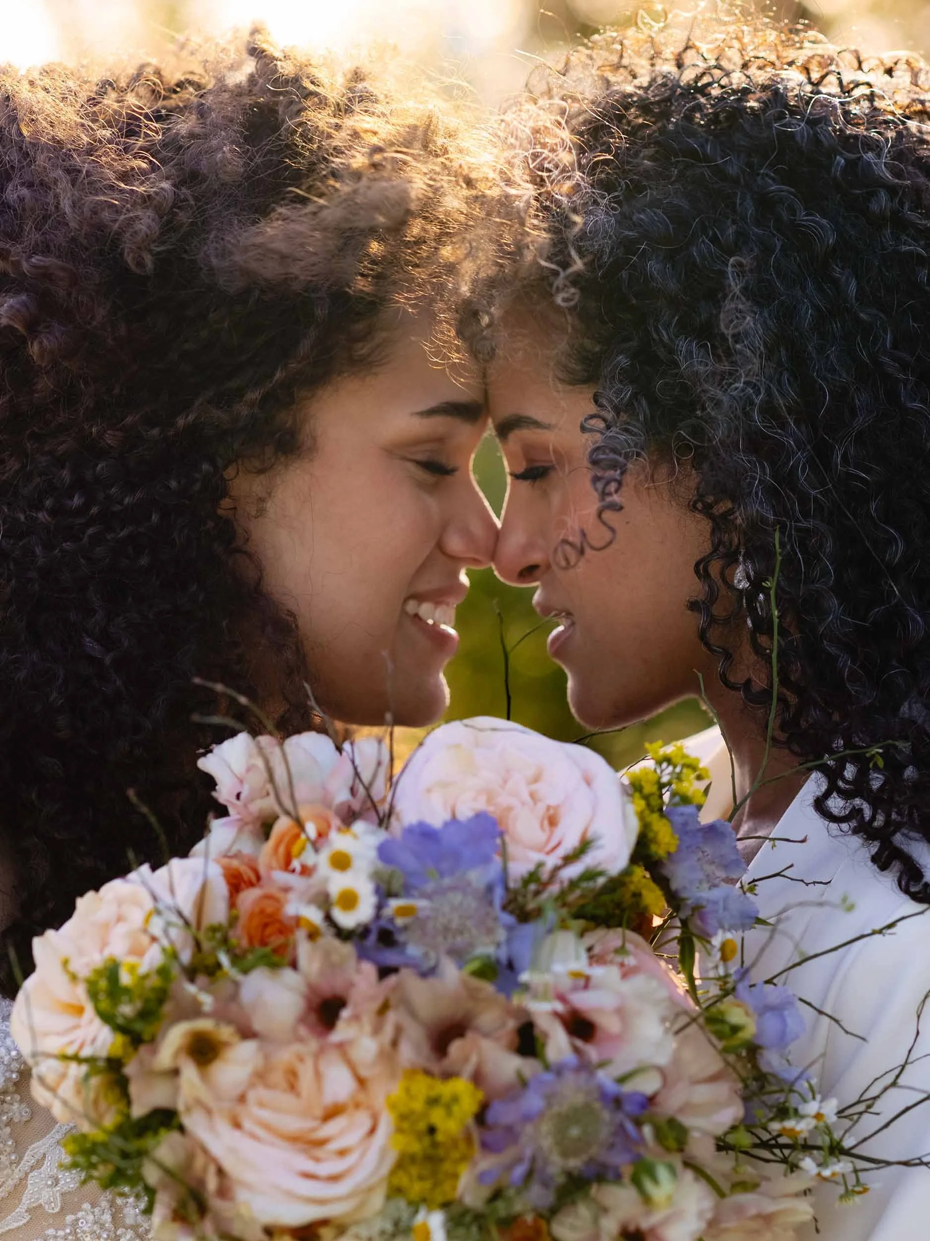 LGBTQ brides smile as they lean in over a colorful bridal bouquet. Sunlight shines through their curly hair at their elopement near Vancouver, WA.