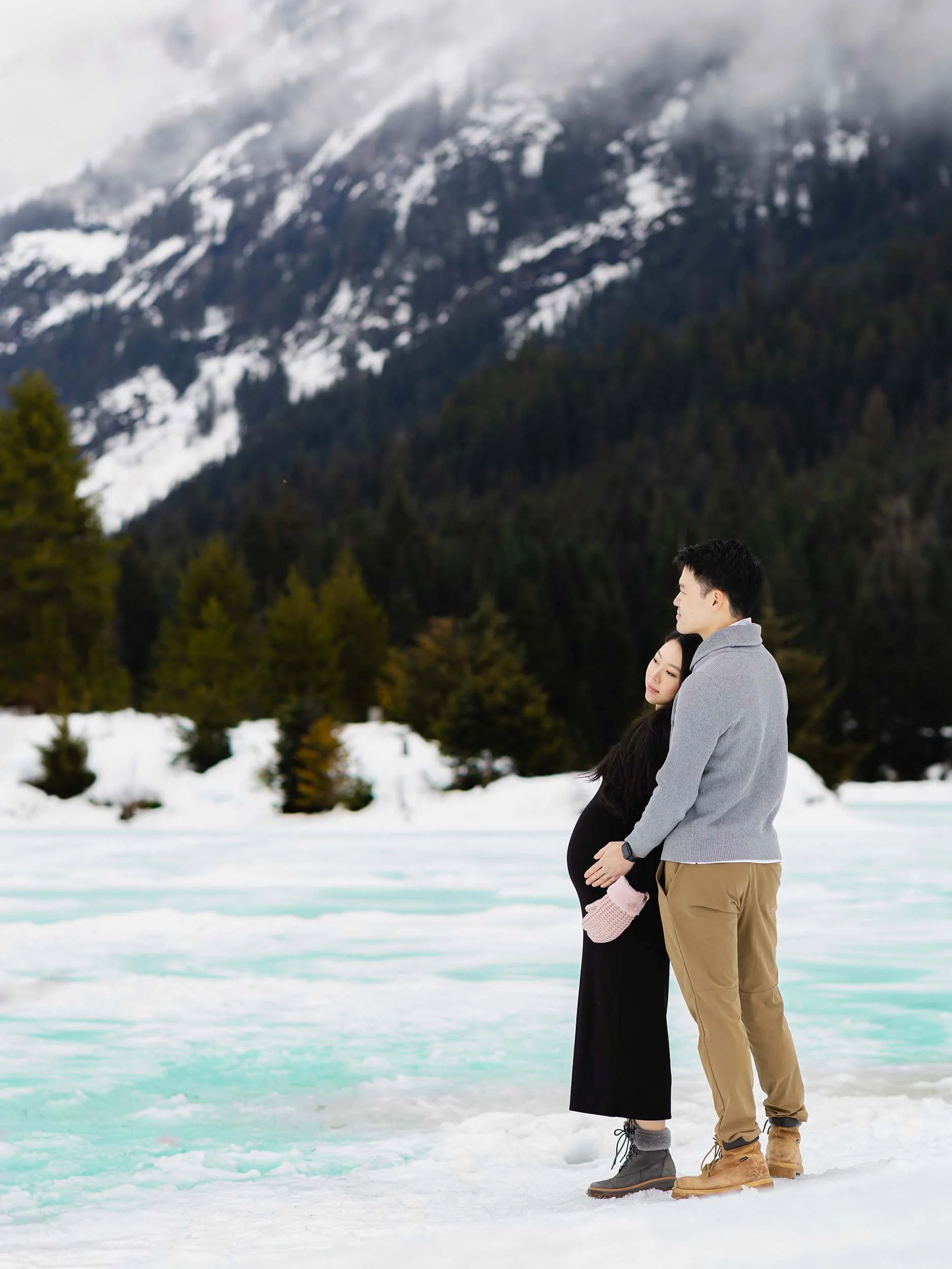 Maternity couple stand at the edge of a frozen, turquoise Gold Creek Pond. Mountains are blanketed in a moody PNW fog. 