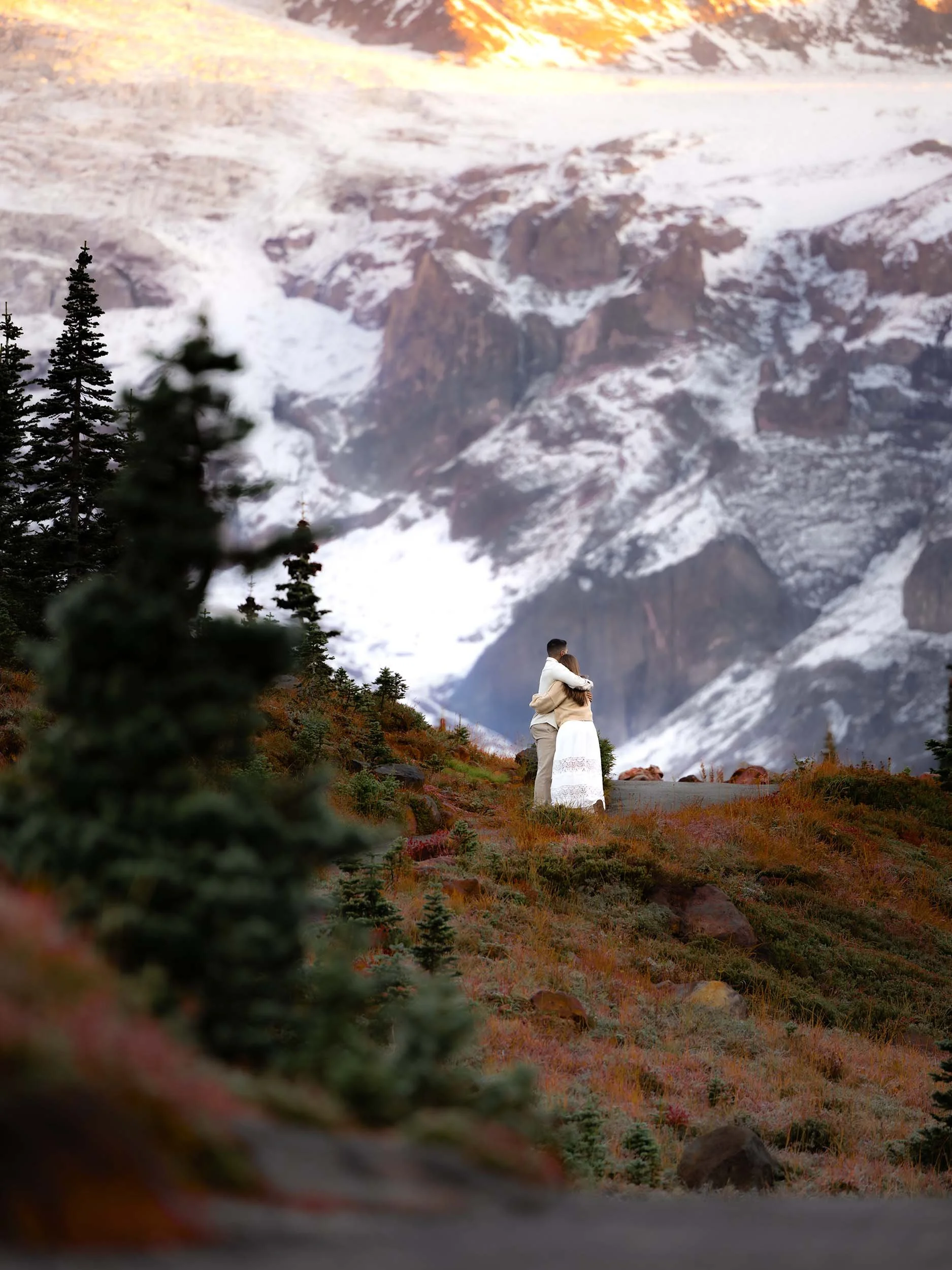 A couple hug, surrounded by fall foliage and a sprawling view of Mount Rainier.