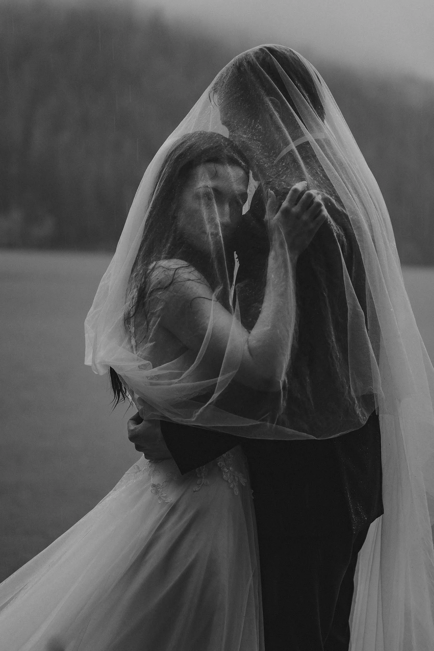 A bride and groom stand under her bridal veil in the rain at Lake Crescent during their elopement. Edited in black and white to enhance the dramatic moment..