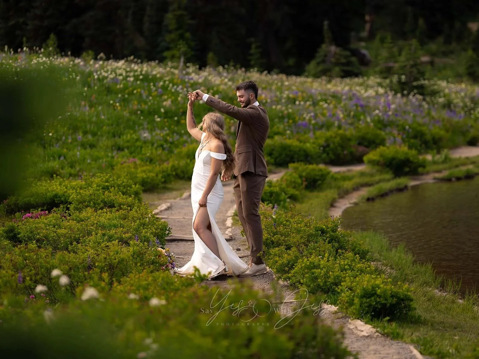 A man in a brown suit twirls his bride, dancing along the path at Tipsoo Lake surrounded by wildflowers on their elopement day.
