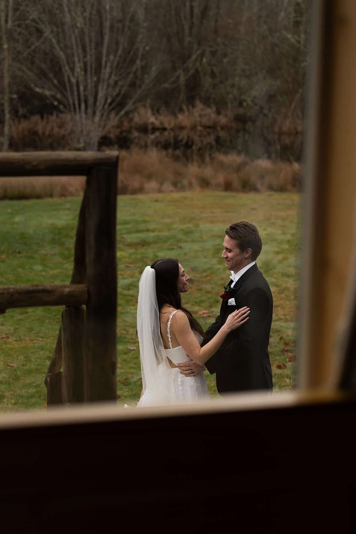 A bride and groom smirk at each other fondly in their wedding attire, as photographed from a perspective from inside the cabin looking out.
