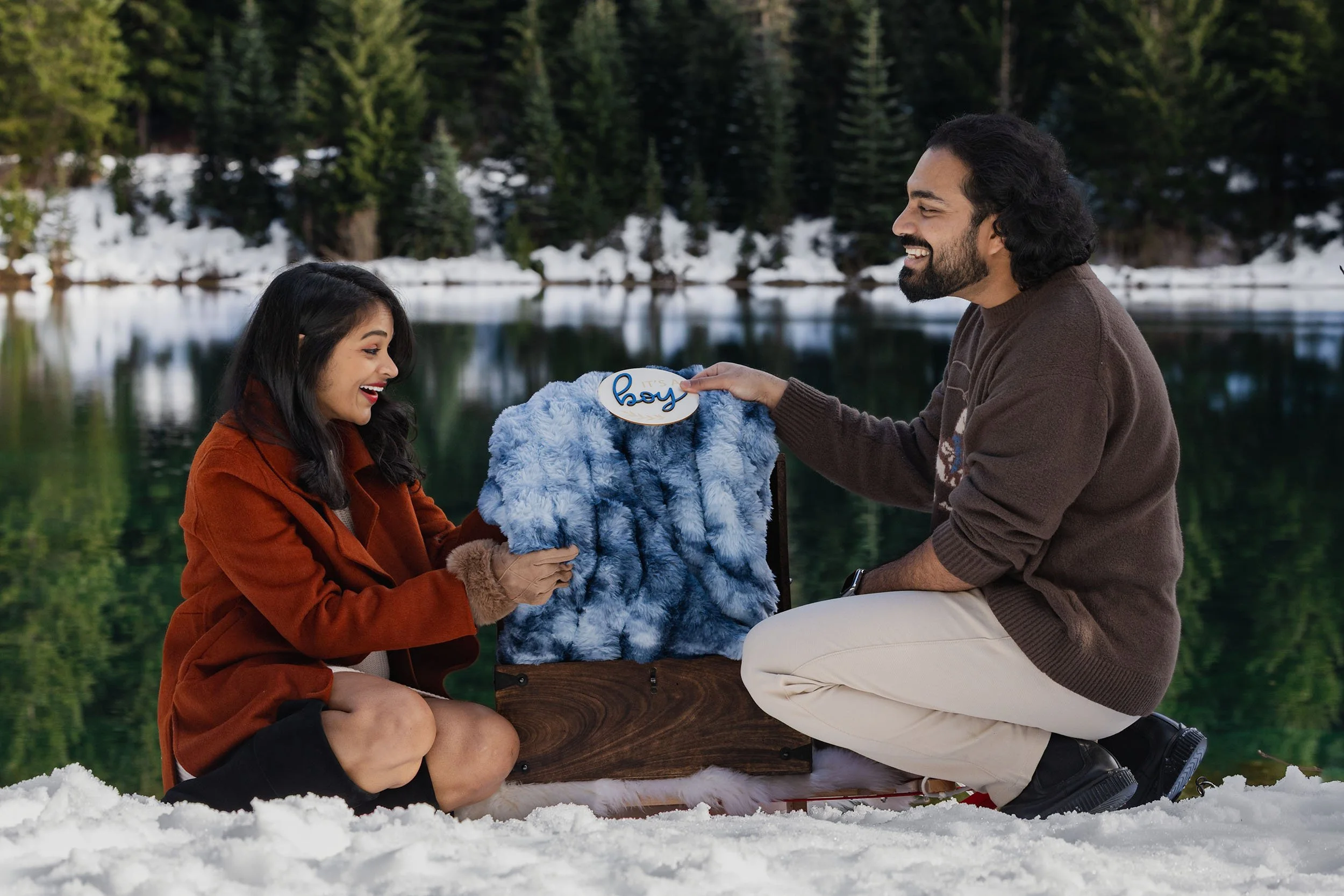 An expecting couple pull a blue blanket and "boy" sign from a box resting on a sled in the snow at a Gold Creek Pond gender reveal.