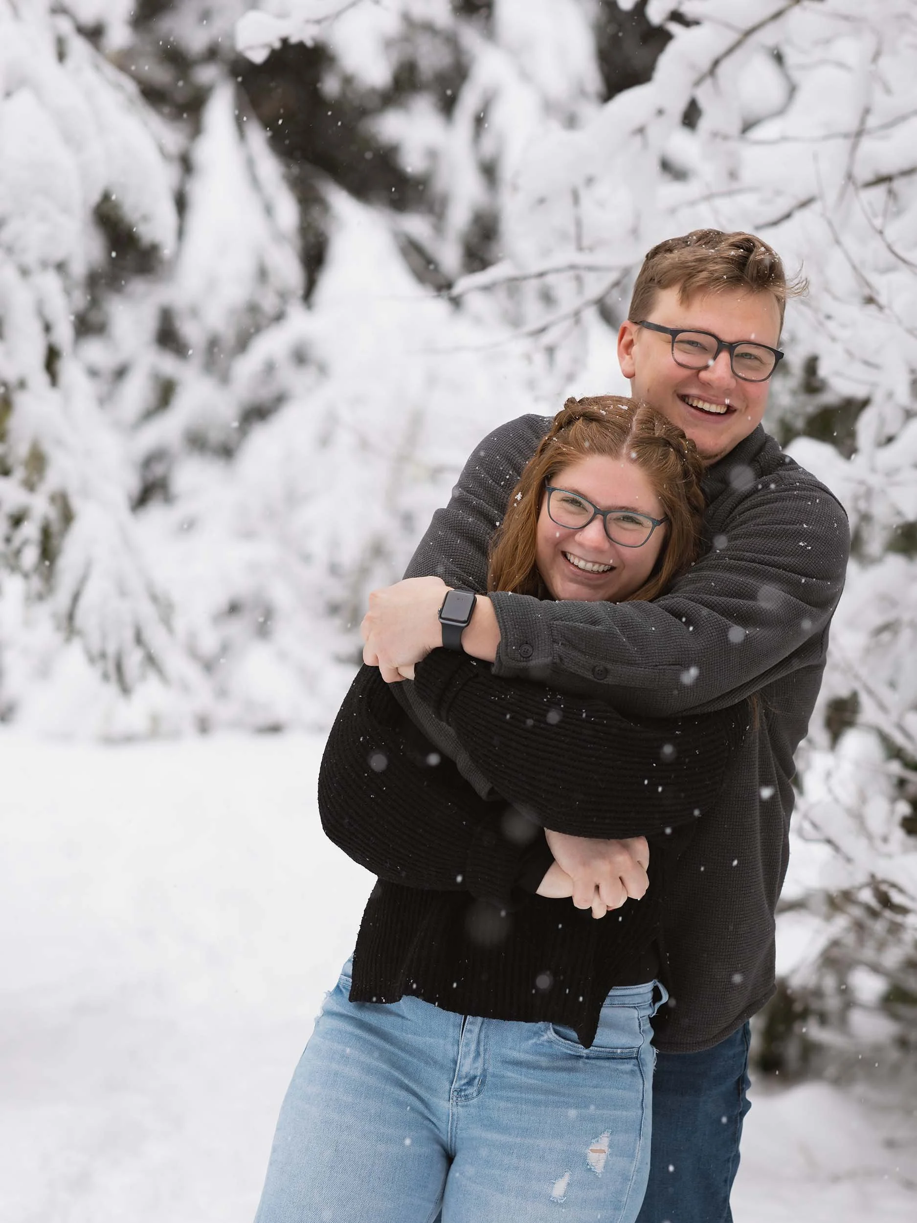 A smiling young couple in winter clothing hugging in a snowy landscape with snow-covered trees during an engagement session in Mount Baker Snoqualmie Forest.