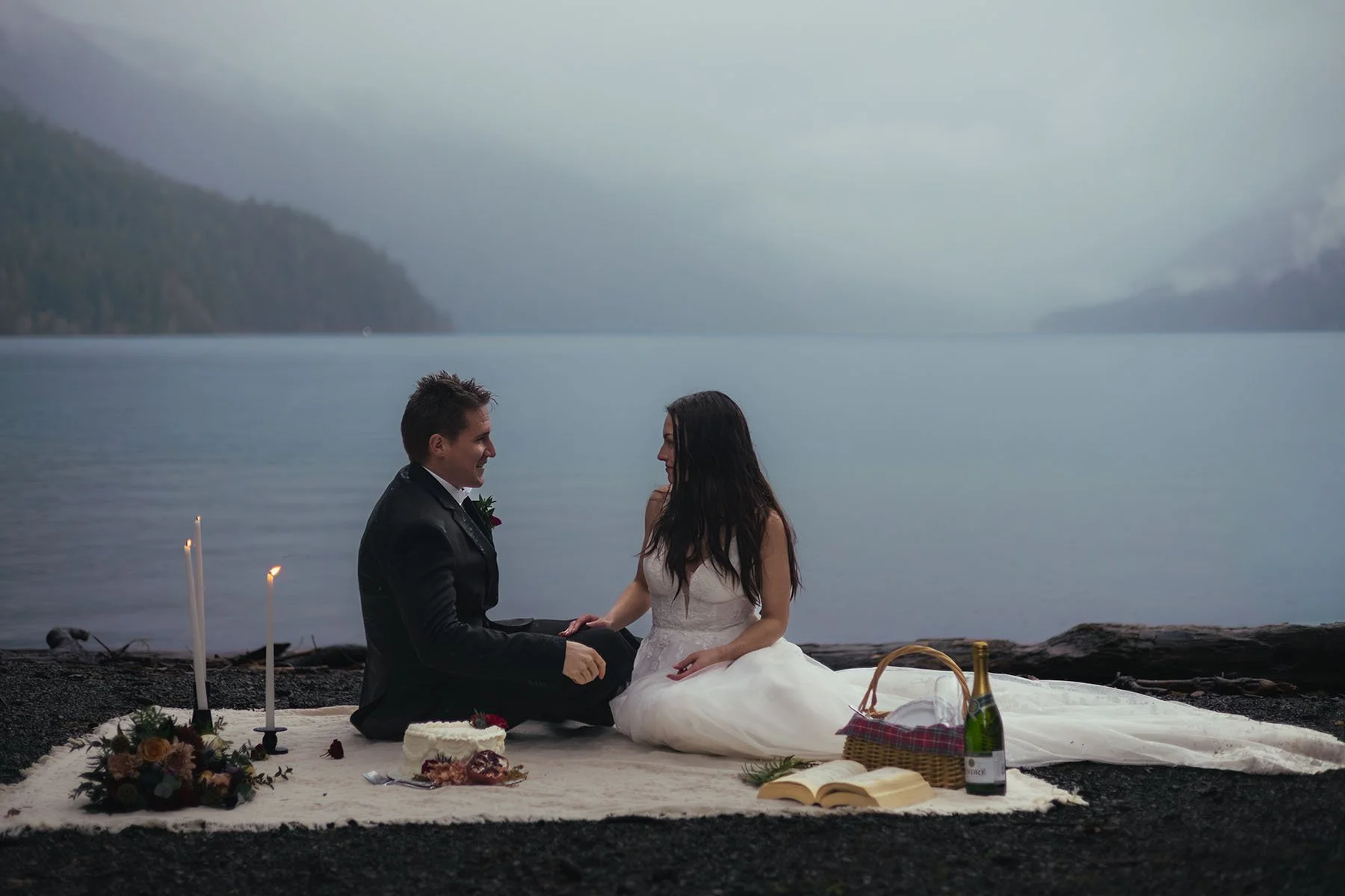 A bride and groom sit on a blanket with candles and wedding cake Lake Crescent during their elopement. The dense fog and blue hour create a dramatic effect.