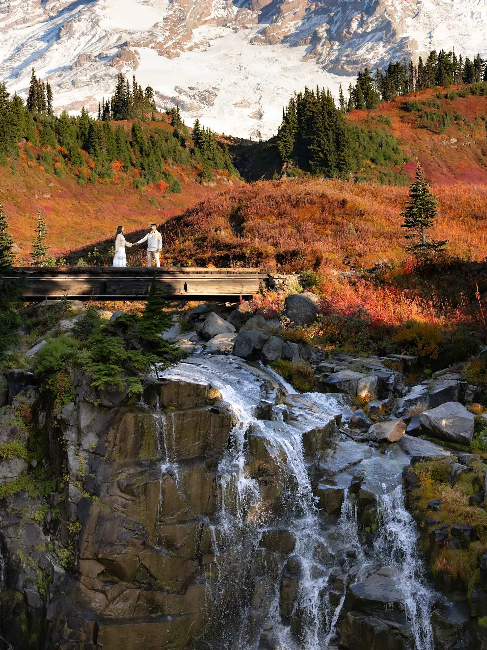 Dramatic view of Mount Rainier with a couple holding hands over Myrtle falls, surrounded by vibrant fall foliage.
